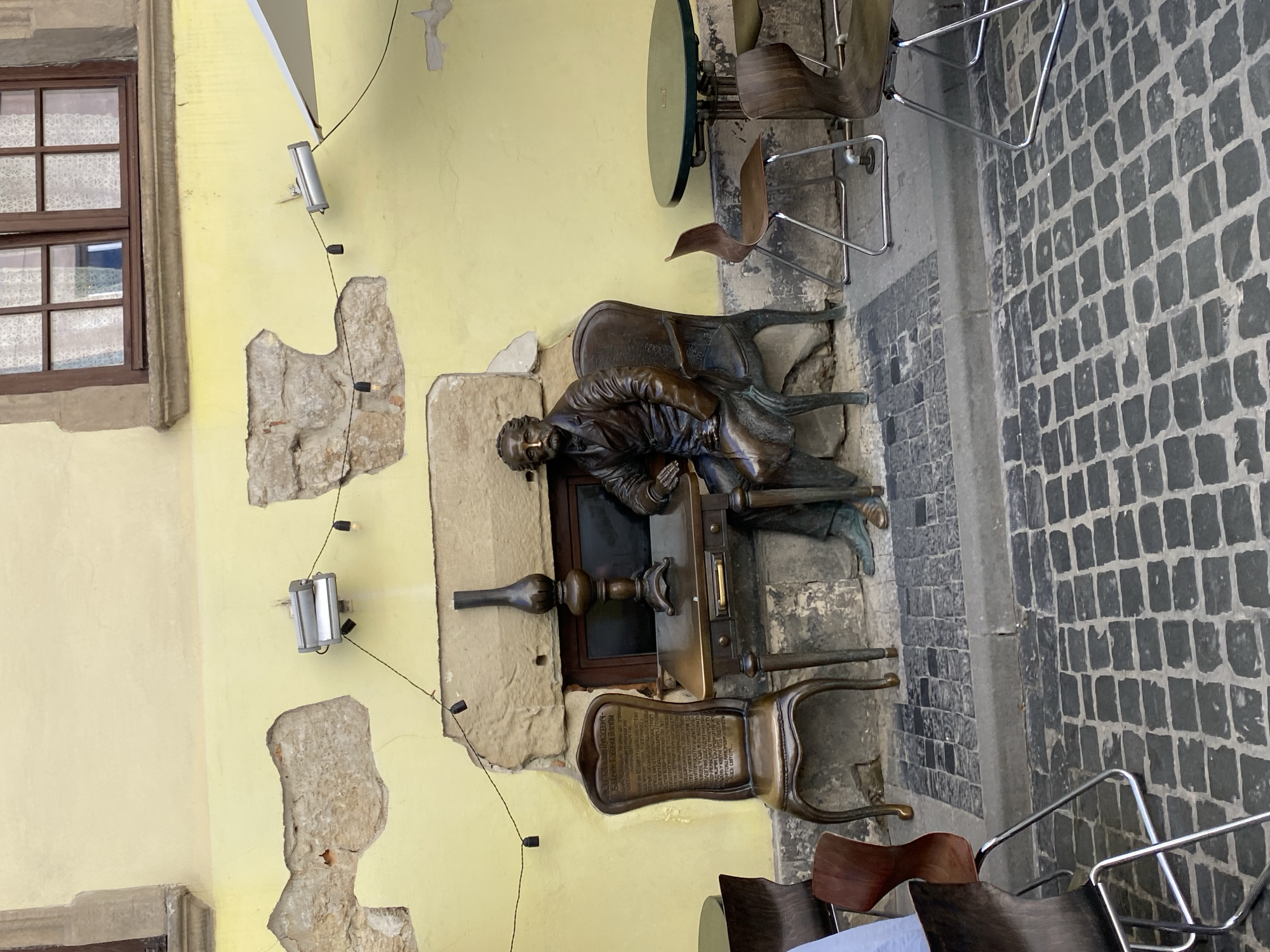 The image depicts a street scene in Lviv, Ukraine, featuring a bronze statue of a man seated at a desk. The statue is placed outdoors, suggesting a public space, possibly a cafe or a square. The statue is positioned in front of a yellow wall with rough, uneven plaster and exposed brickwork, indicating an older building. Above the statue, a window with a dark wooden frame and patterned curtains is visible.
The statue itself is detailed, showing a man in formal attire, with a mustache, wearing a suit and tie. He is seated in a chair at a desk, on which rests an antique lamp. To the left of the desk, a bronze chair with an inscription on its back is placed. The floor is paved with dark cobblestones. In the background, several outdoor cafe tables and chairs are visible, including a round green table and wooden chairs with metal legs. String lights are strung across the scene, attached to the building. The lighting suggests it is daytime, with diffused sunlight.