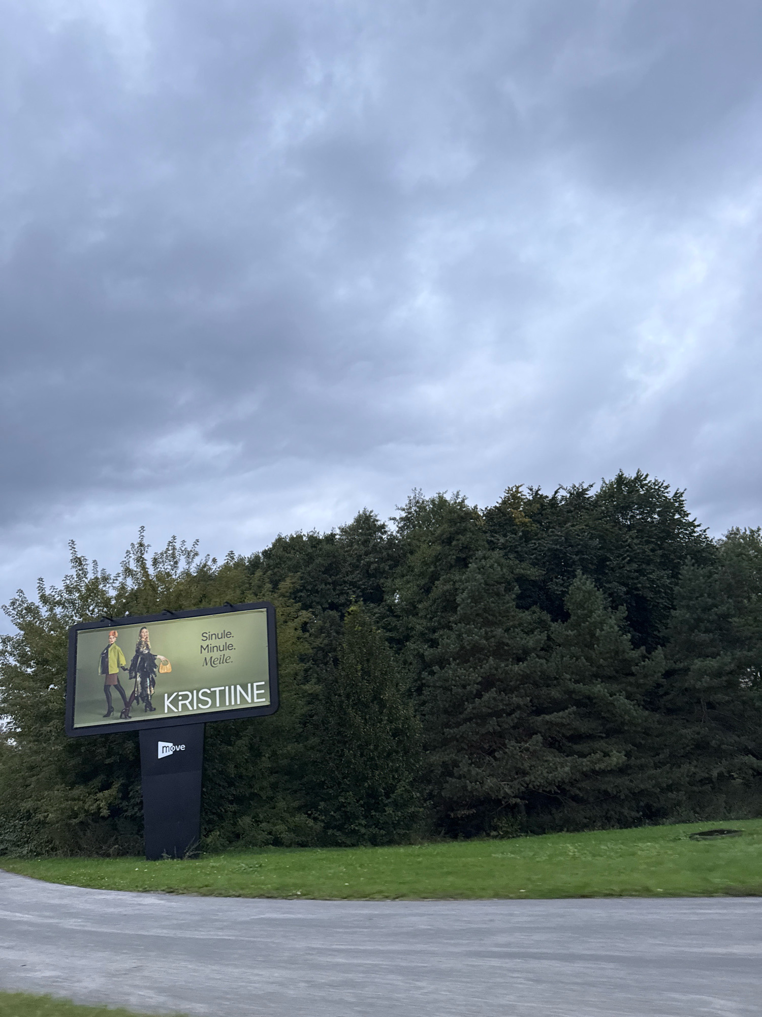 The image shows an outdoor billboard located in Tallinn, Estonia, likely in the Kristiine district, as indicated by the text on the billboard. The billboard features an advertisement for "Kristiine" which appears to be a shopping center or a brand. Two models are depicted on the billboard, showcasing fashion items, possibly for a seasonal campaign. The text "Sinule. Minule. Meile." which translates to "For you. For me. For us." is also visible. Below the advertisement, the logo "move" is displayed. The billboard is situated amidst lush green trees and foliage, with a grey, overcast sky overhead. A paved road or pathway is visible in the foreground. The overall scene suggests an outdoor advertising space in a semi-urban or suburban environment.