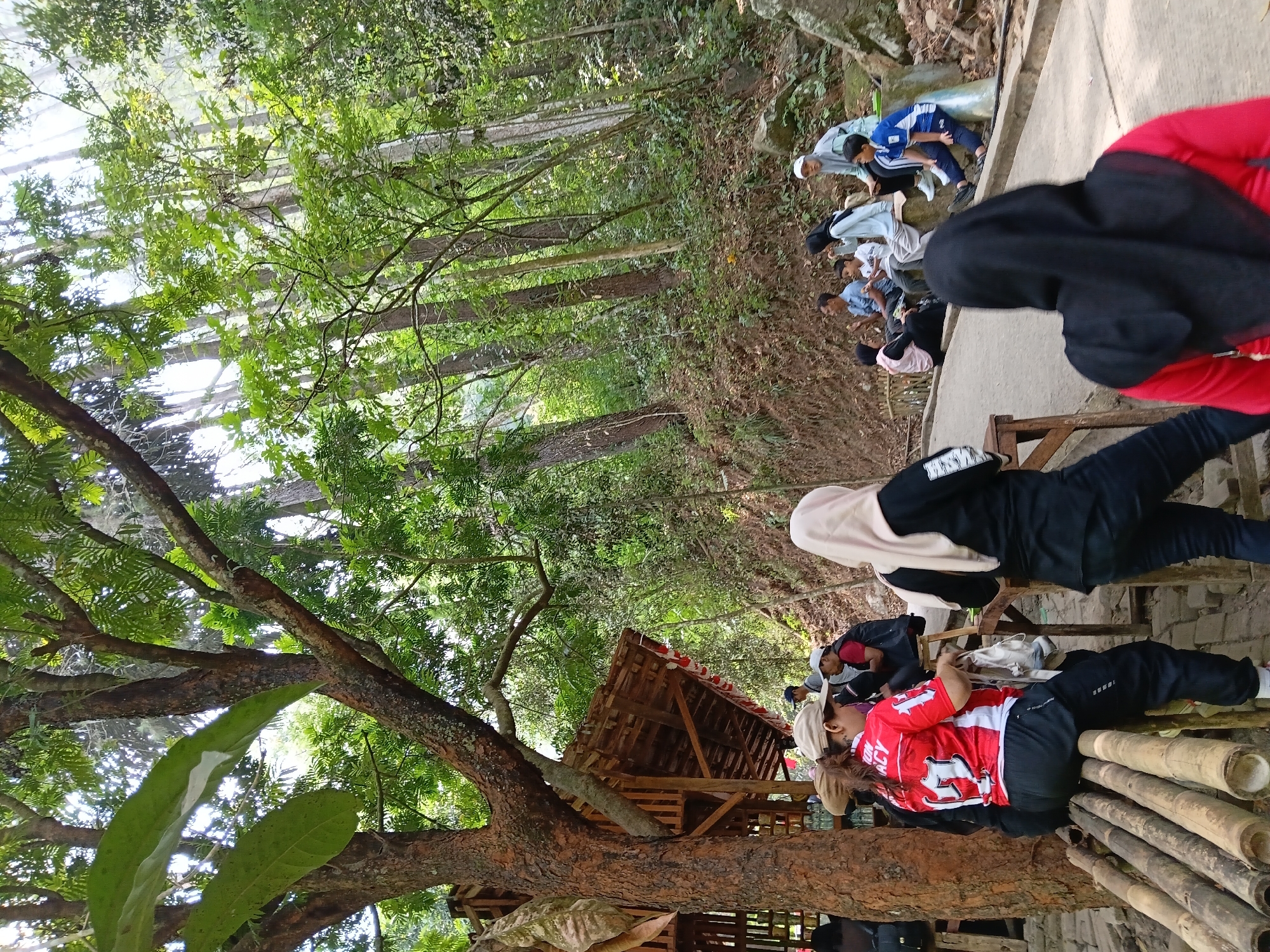 A group of people are gathered in a wooded area, possibly a park or a nature reserve. In the foreground, a woman wearing a red and white jersey with the number 17 and black track pants is seated on a bamboo bench. Next to her, partially visible, is another person wearing a black hijab and black pants. Further back, a group of individuals are seated on a concrete ledge and the ground. The environment is characterized by lush green trees, a dirt path, and rocky terrain. To the left, a wooden structure resembling a shelter or a small building with a thatched roof is visible. The overall atmosphere suggests a casual outdoor gathering. The scene is likely located in Ciburial, Indonesia, given the context of the surrounding environment and vegetation typical of such regions.
