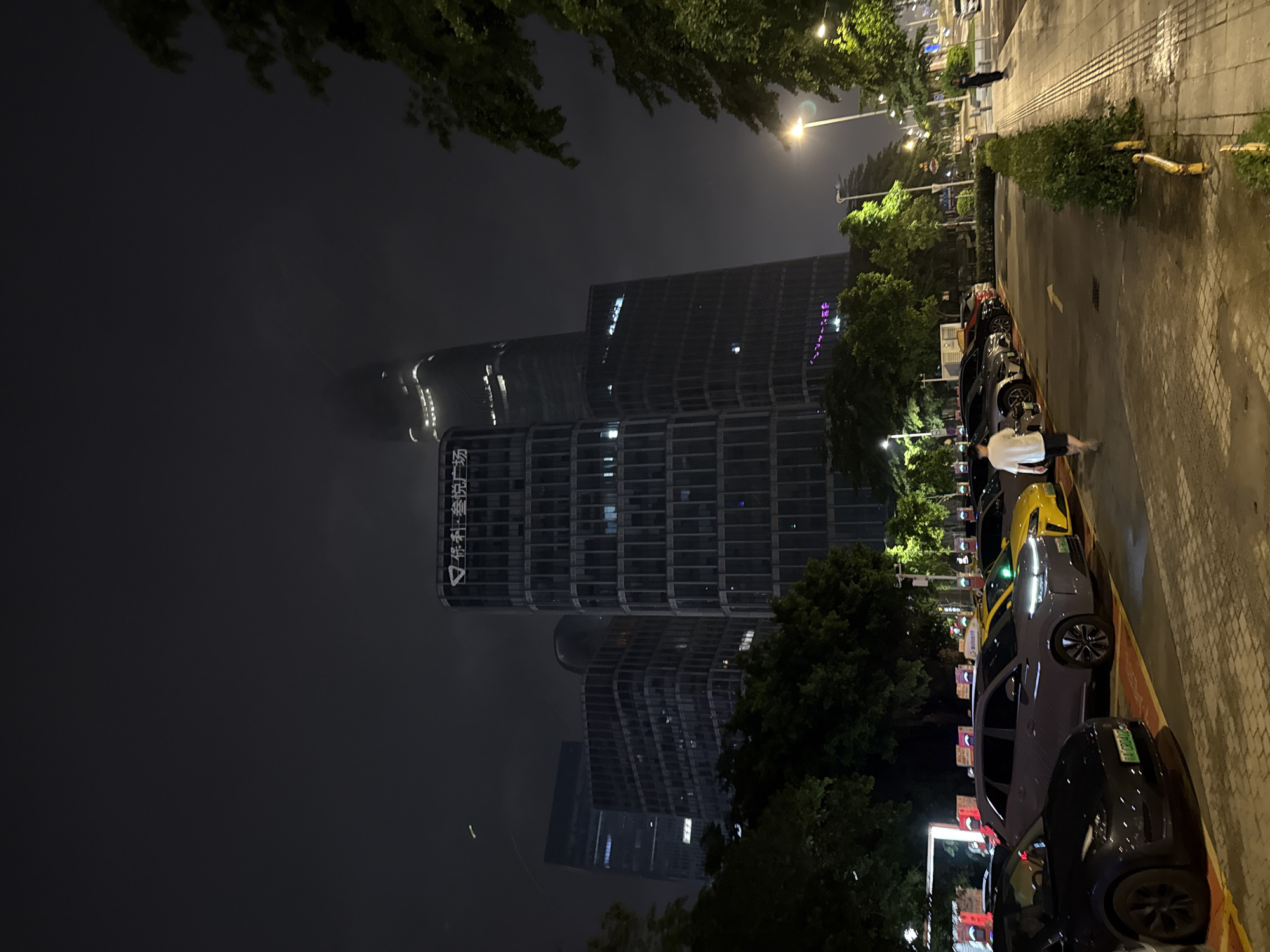 In Haizhu District, China, at night, a row of parked cars is visible in a paved lot. The cars are mostly dark-colored, with a grey SUV and a bright yellow sedan among them. A person in a white t-shirt and dark shorts walks away from the vehicles towards the right side of the frame. In the background, several modern skyscrapers dominate the skyline, illuminated against the dark, foggy sky. Trees line the left side of the parking lot, and streetlights and pedestrian walkways are present on the right. A sign on one of the buildings reads "保利·誉湾广场" (Poly·Yuwan Plaza).