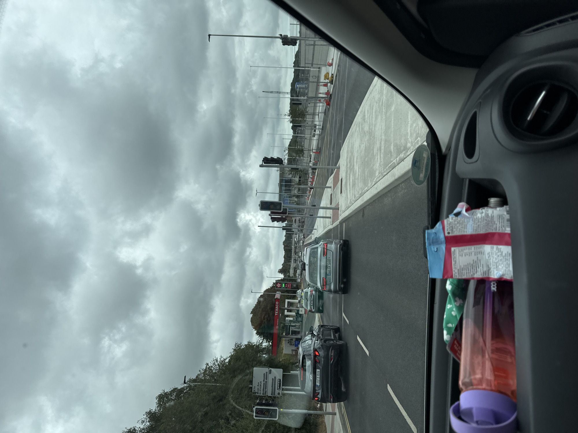 The image depicts a scene from inside a vehicle, looking out onto a road in Waterford, Ireland. The sky is overcast with heavy clouds. In the foreground, on the dashboard, are personal items including a water bottle and a packet of snacks.
The road is paved with traffic markings and a concrete sidewalk. Several cars are visible on the road, including a black Hyundai Tucson and a silver Audi A6. Further down the road, a Texaco gas station is visible, with red awnings and signage. Traffic lights are present at an intersection.
To the left of the road, there is a sign indicating route R711, which leads to a bus route and the city center. Trees and vegetation line the left side of the road. On the right side, there is construction work with barriers and traffic cones. In the distance, a construction crane is visible. The overall impression is of a suburban or semi-urban environment, possibly on the outskirts of Waterford.