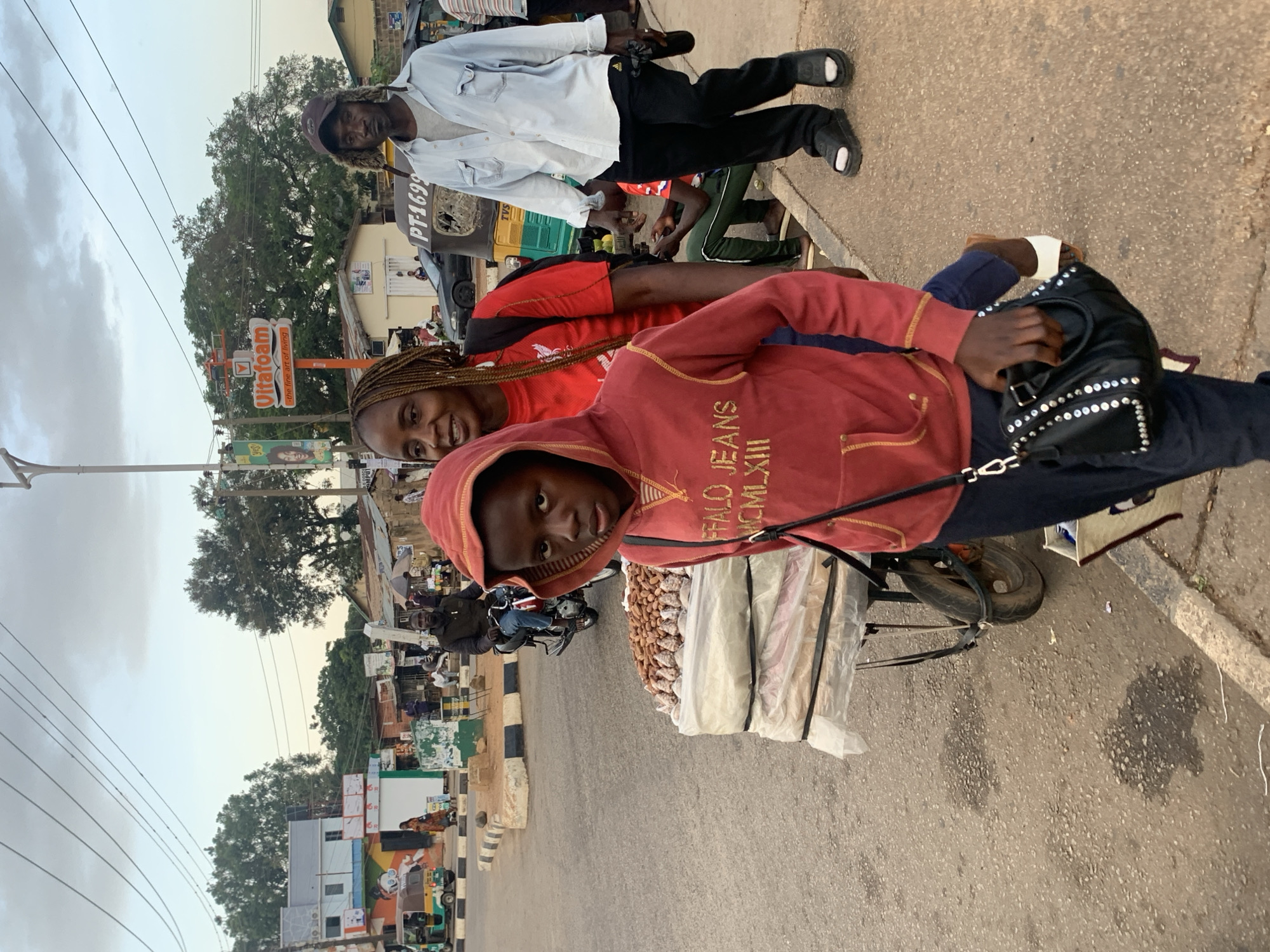 In Barakin Lerawi, Nigeria, an outdoor scene captures a bustling street with pedestrians and vehicles. In the foreground, a young person wearing a red hooded sweatshirt and dark pants walks beside a woman with braided hair, who is wearing a red t-shirt and a black backpack. The young person is carrying a black handbag adorned with silver studs. To their right, a man in a light blue button-up shirt, dark pants, and black sandals walks past. Behind them, a street vendor's cart is visible, piled with goods wrapped in plastic, topped with what appears to be nuts. In the background, a man rides a motorcycle, and various buildings and trees line the street. A sign for "Vitafoam - the fine art of living" is prominent. The overall atmosphere suggests a lively urban environment during daylight hours.