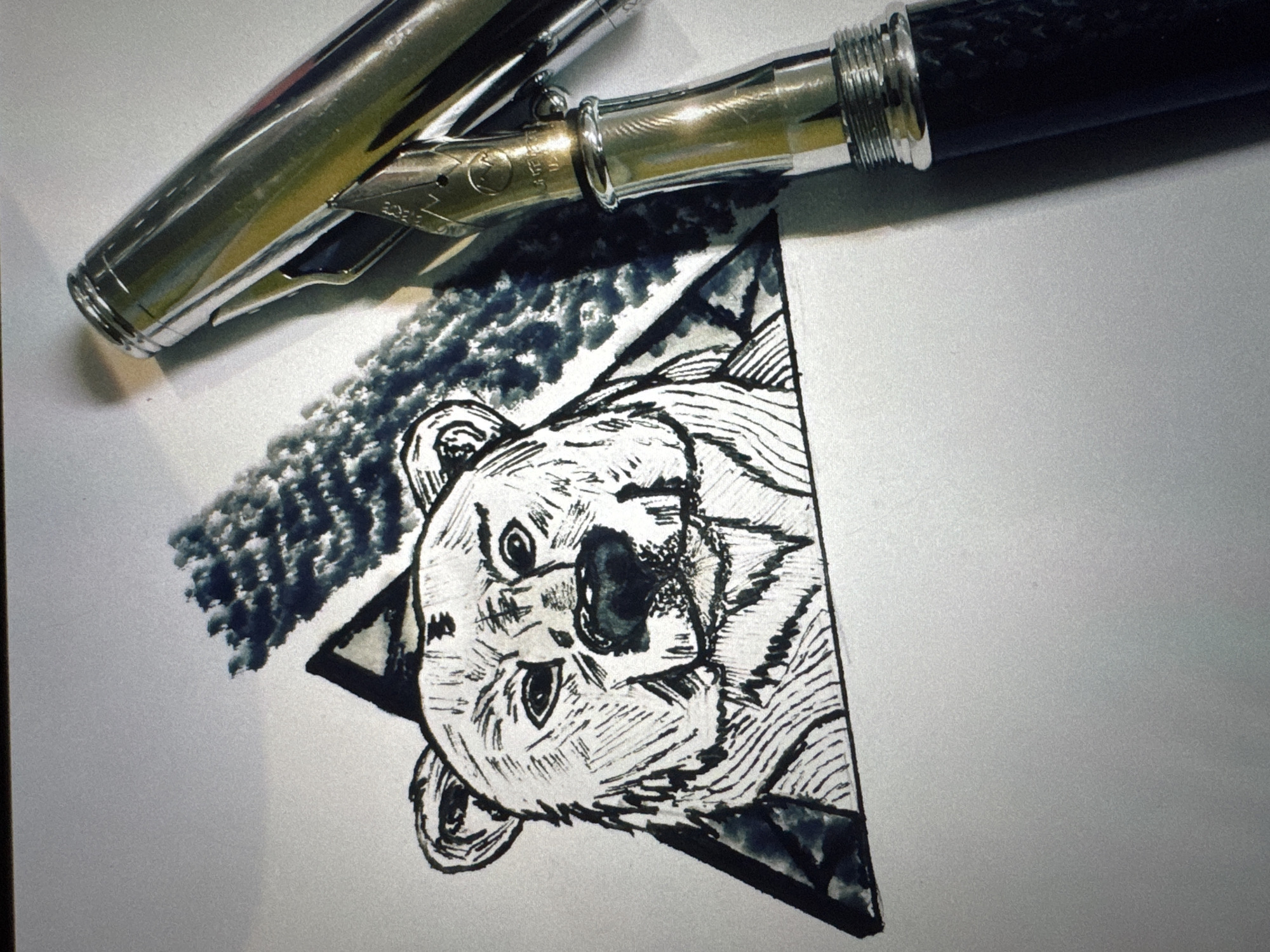A close-up, overhead shot shows a sketch of a bear's face within a triangle, rendered in black ink on white paper. A fountain pen with a silver nib and dark grip rests on the paper, partially obscuring the drawing. The bear's features are depicted with bold, expressive lines, and a textured, splotchy shadow is drawn behind its head. The background is a plain white surface, suggesting a desk or table. The image is devoid of any geographical markers and therefore cannot be associated with Riga, Latvia.