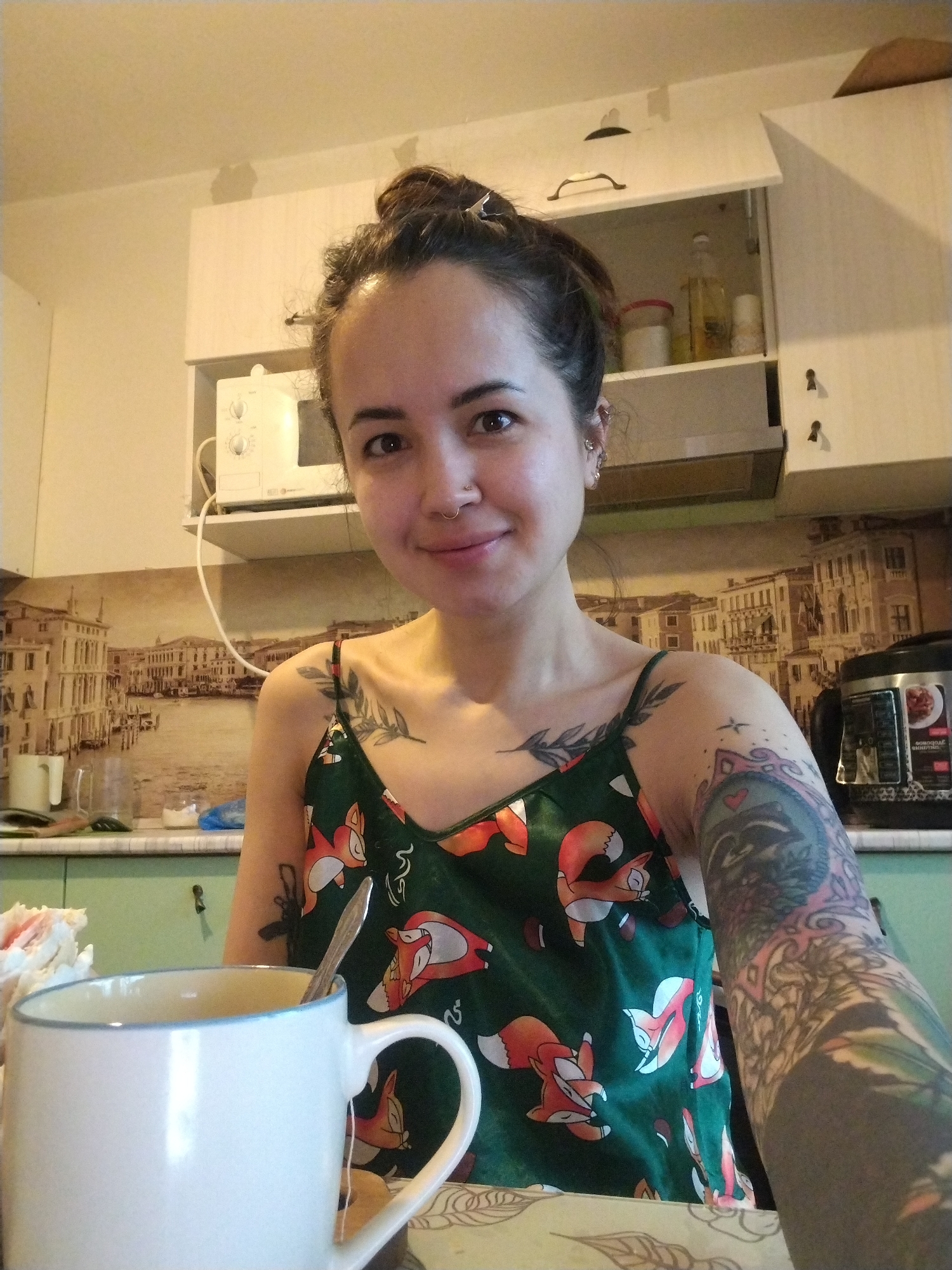 A young woman is captured in a close-up, chest-up selfie or portrait, smiling softly directly at the camera. She appears to be in a kitchen setting in Noviy, Russia, indicated by the background and visible text. Her dark hair is pulled back in a bun, and she has dark eyes and a nose piercing. She wears a dark green spaghetti-strap top adorned with a pattern of orange foxes.
Visible tattoos include a leafy branch design on her left shoulder/collarbone area and a detailed, colorful sleeve tattoo covering her entire left arm. The sleeve features various elements, including what appears to be an animal face (possibly a raccoon or similar creature) with a heart above it, and intricate swirling patterns.
In the foreground, a white mug with a light blue rim contains a tea bag and a spoon, suggesting she is having tea or coffee.
The background reveals a typical kitchen environment. A white microwave oven rests on a shelf above a counter, beneath white kitchen cabinets. One cabinet door is ajar, revealing bottles and containers inside. The backsplash behind the counter features a large, sepia-toned image depicting a European cityscape, reminiscent of Venice, with canals and historical buildings. To the right, on the counter, a silver and black multi-cooker or pressure cooker is visible. Text on the multi-cooker, in Cyrillic, includes "МЕНЮ" (MENU) and other program options like "Овсяная каша" (Oatmeal), "Суп" (Soup), and "ПЛОВ" (Plov). The overall lighting suggests it is daytime.