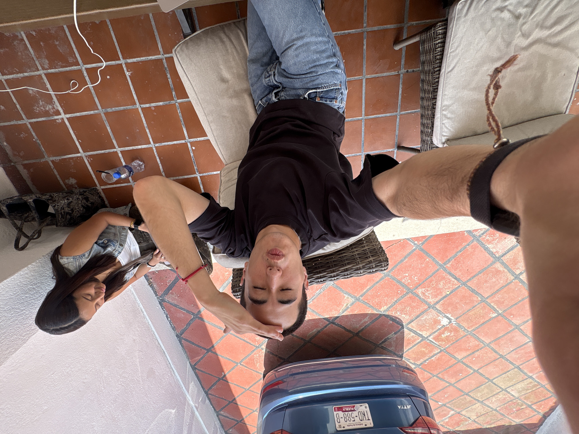 The image captures a man lying on a patio chair with a woman seated nearby. They are positioned next to a blue car, its Puebla license plate visible. The setting appears to be in Puebla, Mexico, as indicated by the plate. The patio is tiled with red bricks, and furniture is made of wicker. The man is in jeans and a dark t-shirt, while the woman wears a denim jacket.