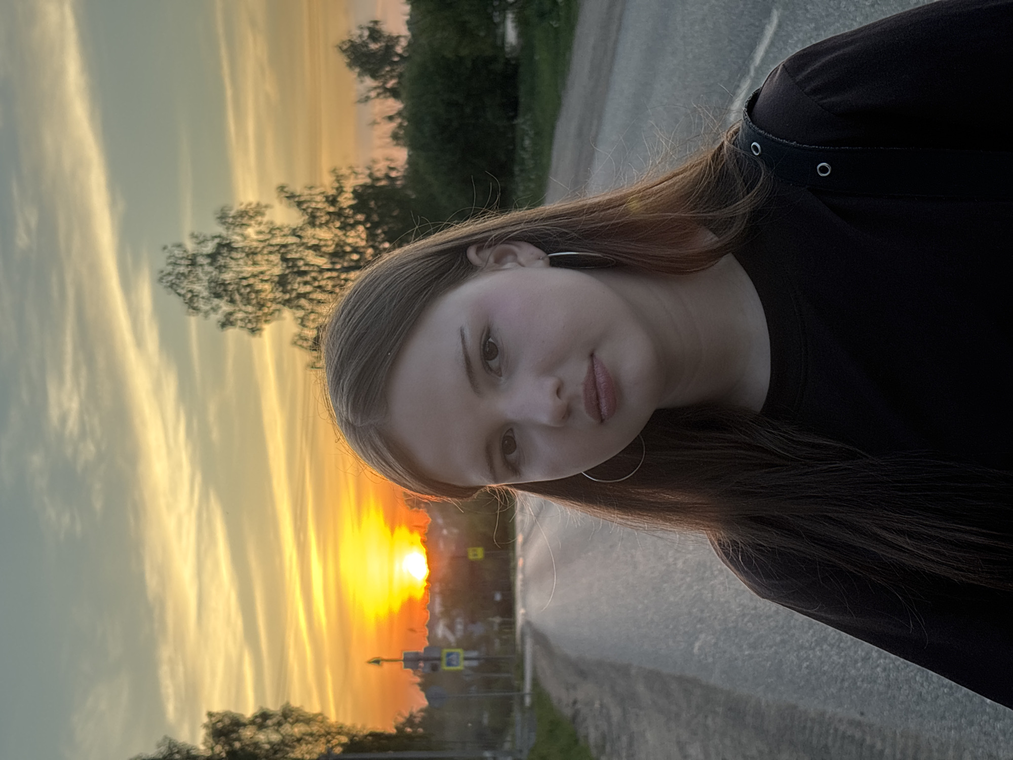 A young woman is posing for a photo during sunset on a road in what appears to be Фоки, Russia. The woman is wearing a black top and silver hoop earrings. The sunset creates a warm, yellow and orange glow, backlighting the woman. The road has green patches of grass and trees adjacent to it. In the distance, a street sign is visible.
