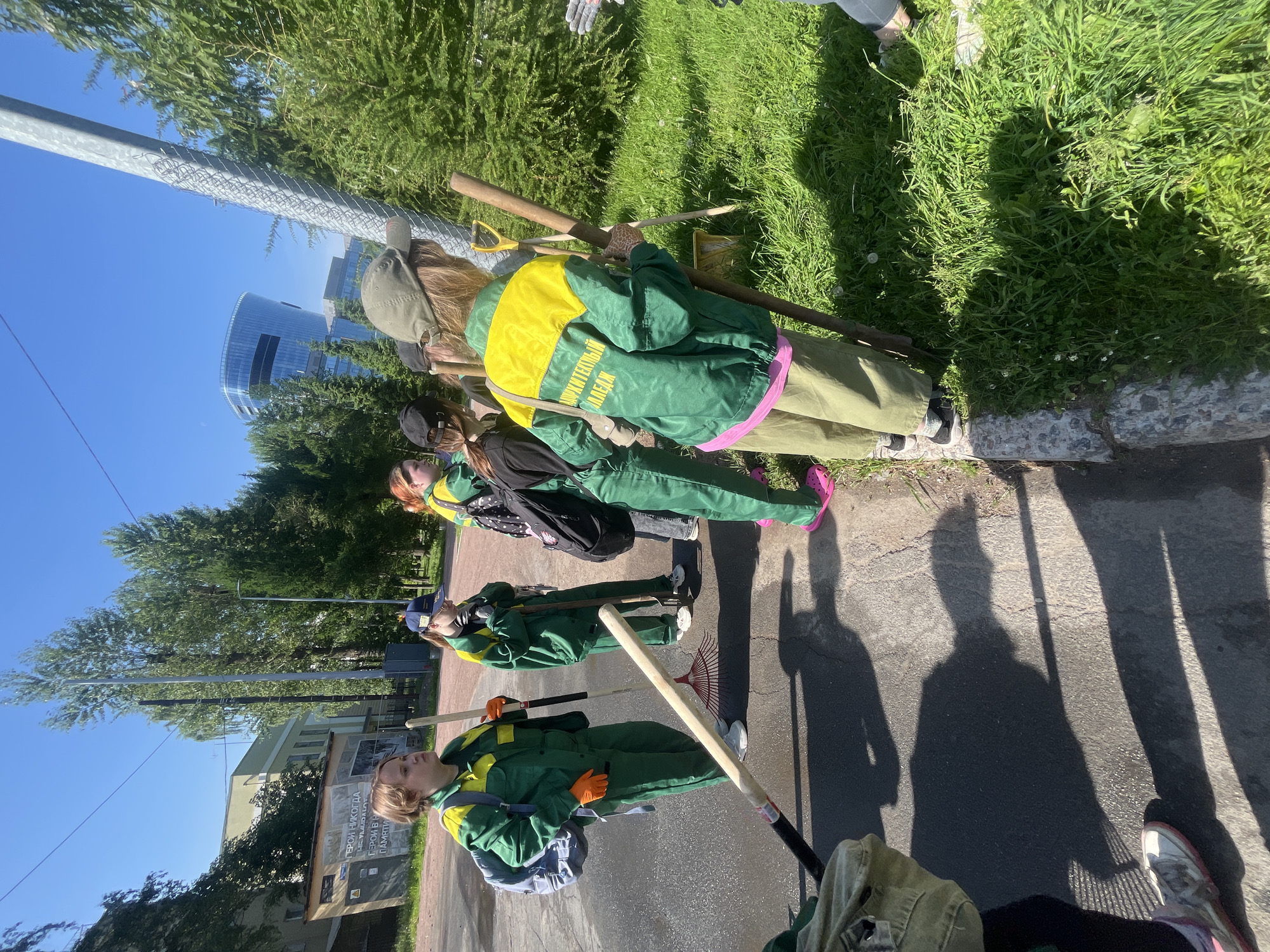 A group of young individuals, likely students or volunteers, are engaged in outdoor work on a sunny day in an urban park or green space in Saint Petersburg, Russia. They are dressed in matching green and yellow work uniforms, suggesting an organized activity like a community cleanup or environmental project.
The individuals are positioned near a paved walkway and a grassy area bordered by a low stone curb. Several people are holding gardening tools such as rakes and shovels, indicating they are likely working on landscaping or tidying the area.
In the background, typical Saint Petersburg urban scenery is visible, including apartment buildings, trees, and a distinctive modern glass building. Power lines and streetlights are also present. The overall atmosphere is bright and active, with the sun casting strong shadows.