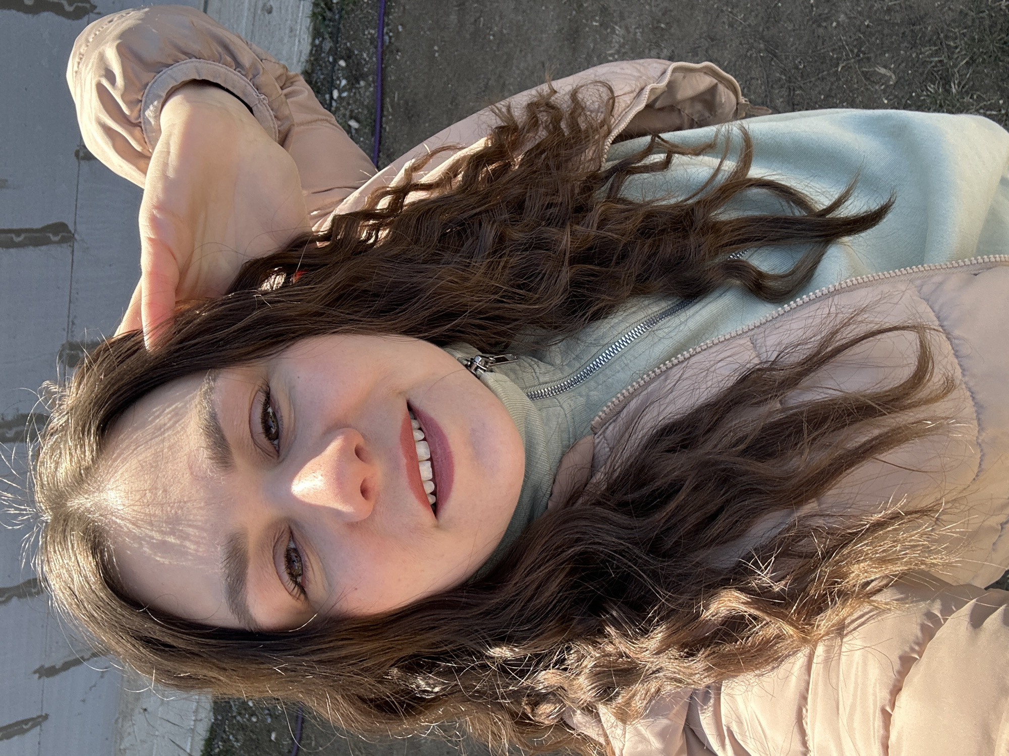 The image depicts a young woman with long, wavy brown hair smiling at the camera. She is wearing a light pink puffer jacket and a light green turtleneck sweater. Her hand is raised near her head. The background appears to be an outdoor setting with concrete and some vegetation visible. The location is Chaikovsky, Russia.