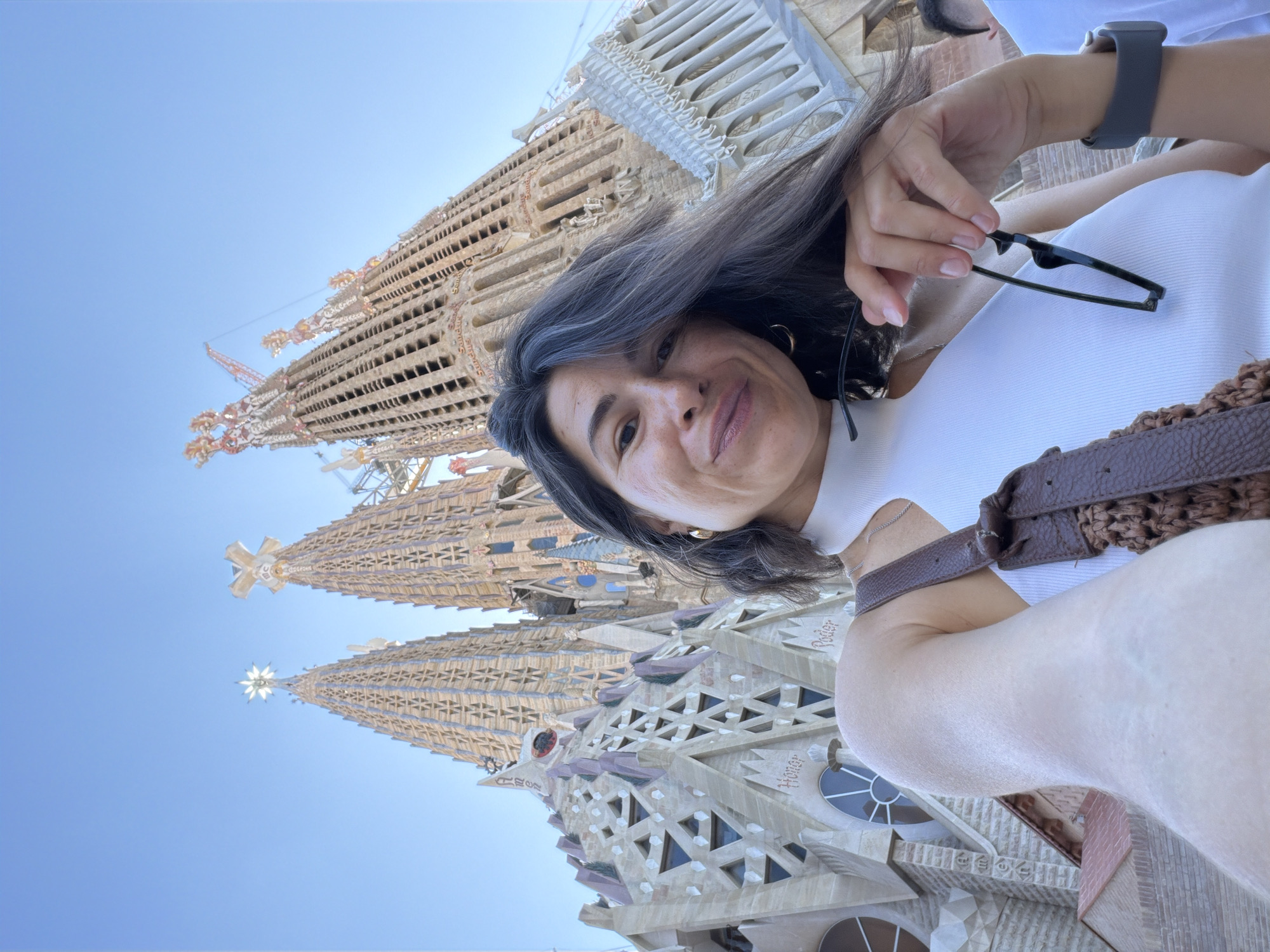 In a selfie taken from a low angle, a woman with dark hair and wearing a white halter top poses in front of the Sagrada Familia in Barcelona, Spain. The iconic basilica, with its intricate spires and stone facade, dominates the background. The woman is holding a pair of black-rimmed sunglasses in her right hand and has a brown crossbody bag slung over her left shoulder. She is looking at the camera with a slight smile. The sky above is clear and blue.