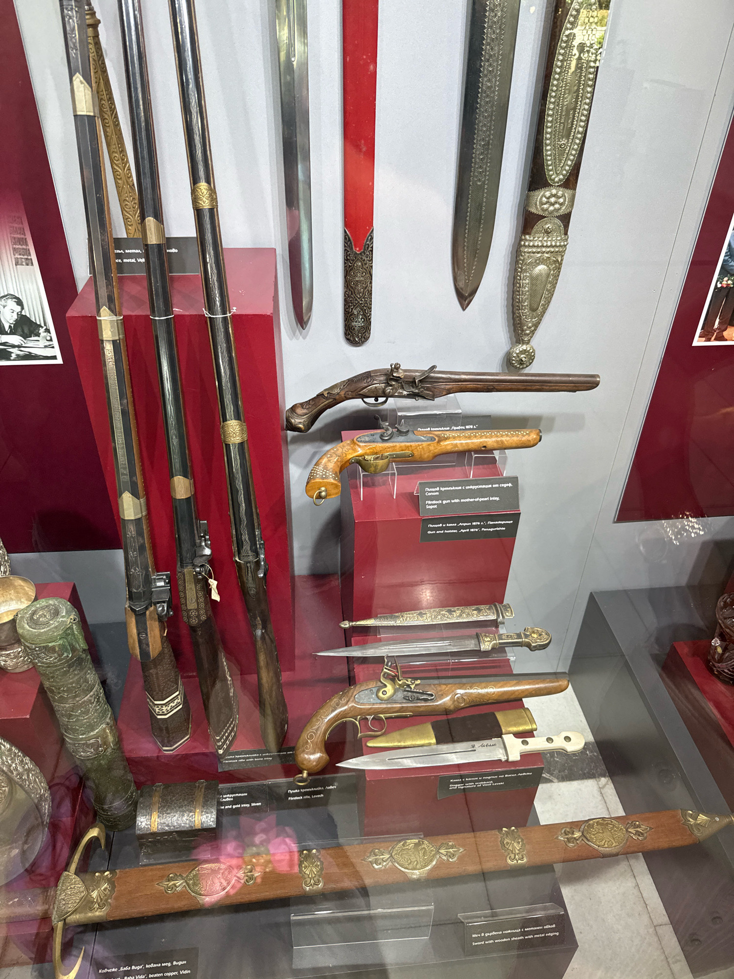 The image displays a museum exhibit featuring a collection of antique weaponry, including rifles, pistols, swords, and daggers. The items are arranged on red pedestals and within display cases, with some items also hung on a grey wall.
In the foreground, a wooden sword sheath adorned with ornate metalwork is visible. To the left of the sword, a metal object resembling a cannon barrel or decorative cylinder is displayed, along with smaller metallic vessels. Several long rifles with decorative metal inlay on their stocks and barrels are positioned vertically on a red pedestal. Above these rifles, two flintlock pistols are showcased. One pistol is longer and darker, while the other has a lighter wooden stock with intricate dot detailing. Below the pistols, several daggers with ornate handles and sheaths are displayed horizontally.
Further up on the grey wall, two sheathed swords are mounted. The sword on the right has a dark wooden sheath decorated with extensive silver metalwork, including intricate patterns and floral motifs. The sword on the left appears to be a straight blade with a decorative scabbard.
The background shows a grey wall and a red display case. A framed photograph of a man in a suit is visible on the far left, and a red object, possibly part of another exhibit, is seen on the far right.
The textual labels in the image provide information about the objects, such as "Flintlock rifle with bone inlay," "Flintlock gun with mother-of-pearl inlay," and "Gun and holster," along with dates and origins.
This exhibit is located in the Ivan Vazov House-Museum in the city of Pravets, Bulgaria. The museum showcases the life and works of the Bulgarian poet Ivan Vazov.