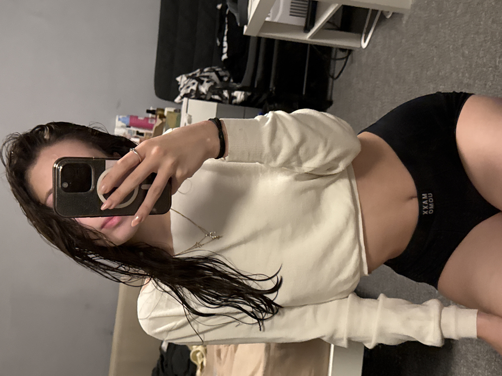 A woman is taking a selfie in what appears to be a bedroom. She is wearing a long-sleeved white cropped shirt and black shorts or underwear. Her hair is dark and wet. The background shows a bed, a black chair, and a white shelf unit. The location in the city of Sofia, Bulgaria, cannot be determined from the image.