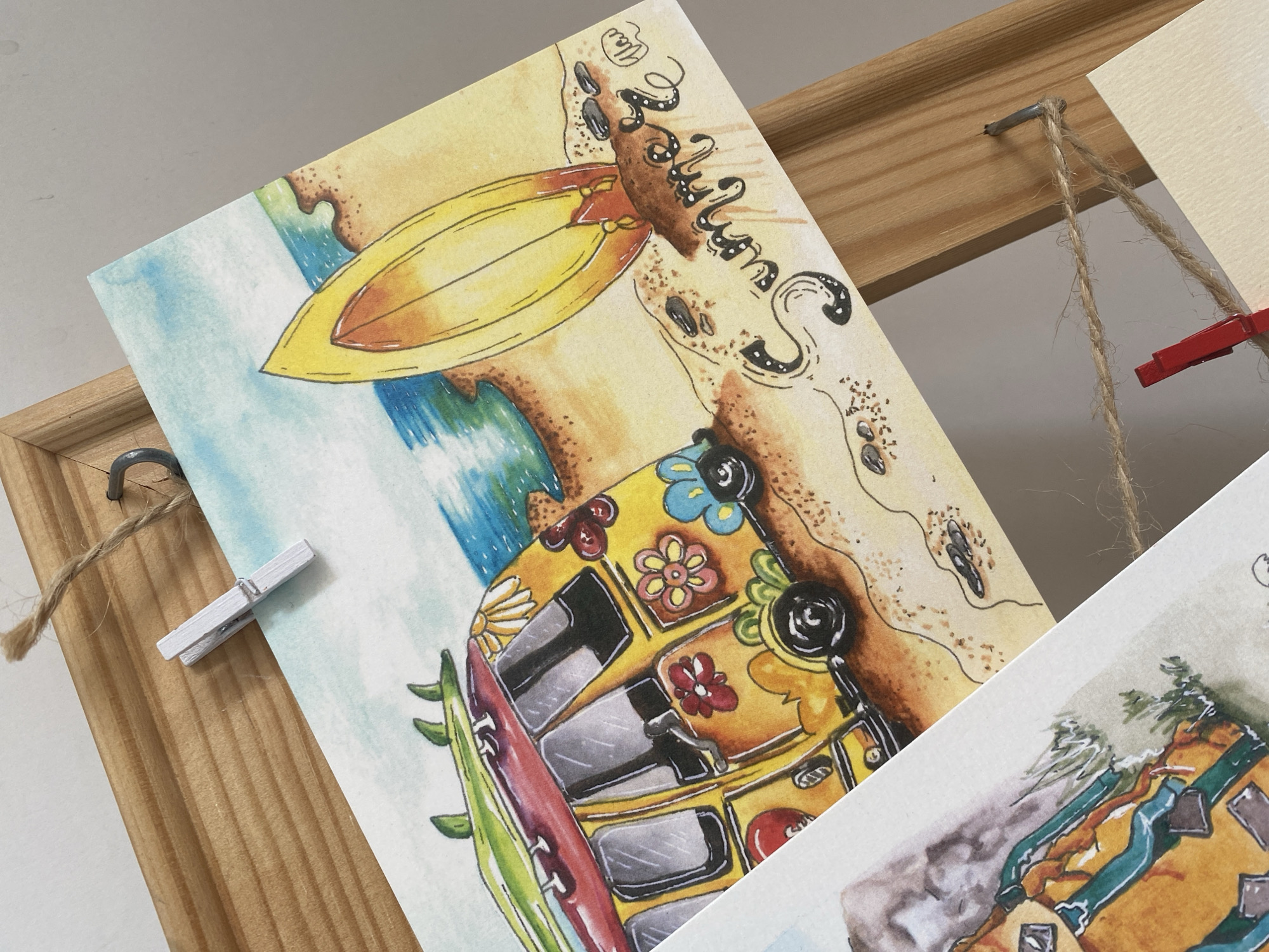 The image shows two watercolor postcards displayed on a wooden frame using twine and clothespins. The postcard in the foreground is angled towards the camera and features a yellow van with flower decorations, a surfboard, and beach scene. The other postcard is partially visible. The postcards are suspended from a wooden frame with twine attached with small nails. One clothespin is white, and another is red. The background is a plain, light surface. The location is not identifiable as Yekaterinburg, Russia.
