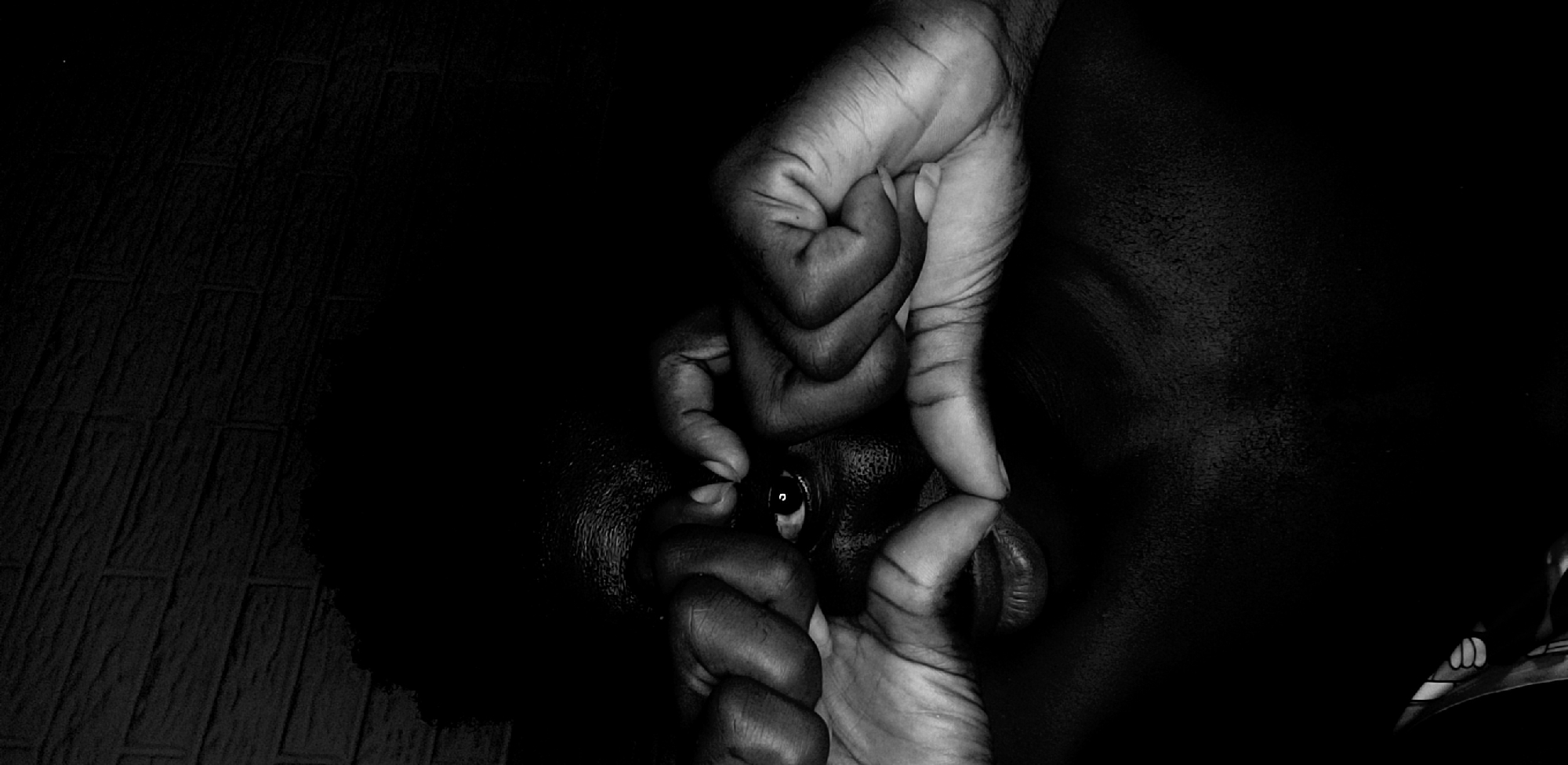 A black and white photograph depicts a human eye and mouth, seemingly obscured by two hands that are positioned directly in front of the face. The hands and the person's skin appear to be dark-skinned. The left side of the image reveals a textured surface, possibly a tiled or patterned wall, while the right side fades into darkness. The setting of this image is in Narayi, Nigeria.