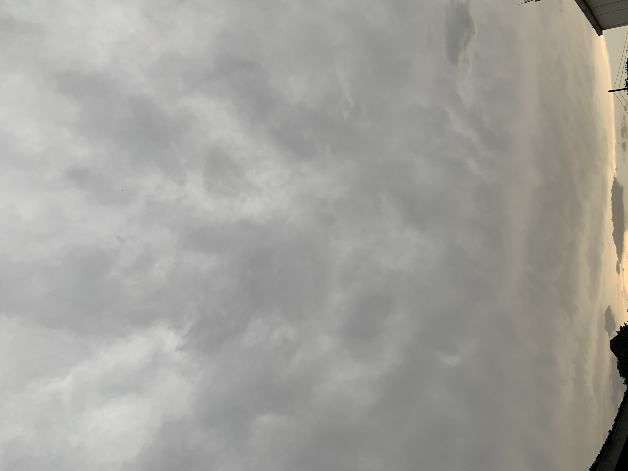The image shows a cloudy sky, predominantly gray. At the top right corner, parts of a building are visible, along with some foliage and power lines.