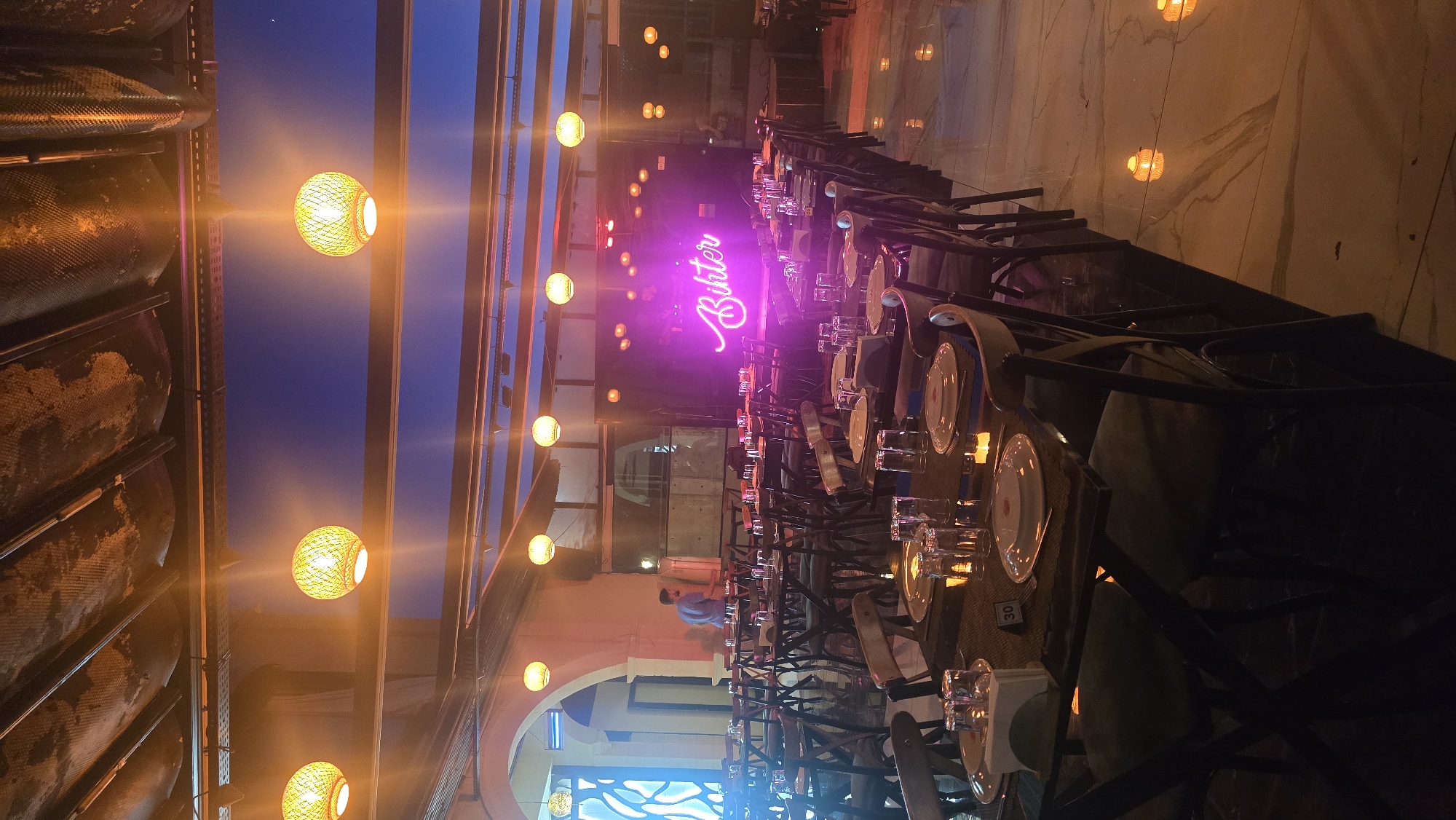The image captures an evening scene within a restaurant or event space, characterized by warm lighting and a lengthy dining setup. Long tables, arranged in rows, are meticulously set with white plates and clear glasses, suggesting readiness for a meal or gathering. The chairs surrounding these tables appear to be a mix of standard dining chairs and possibly bench-style seating.
A dominant feature of the setting is the elaborate lighting. Numerous spherical lamps, crafted from woven material, emit a warm amber glow, casting intricate patterns of light and shadow throughout the room. Through what appears to be a glass ceiling or skylight structure, a dark blue evening sky is visible, reinforcing the time of day.
A vibrant pink neon sign prominently displays the word "Bohter" in a cursive script, serving as a focal point in the background. In the mid-ground, one person wearing a blue shirt is visible, standing near a table and facing away from the camera. The ambiance is inviting and stylish, with a focus on the interplay of light and texture. On one of the tables, a small white tag with the number "30" is visible, likely indicating a table number. The left side of the frame shows a textured wall or partition, possibly made of metal with a patterned surface.