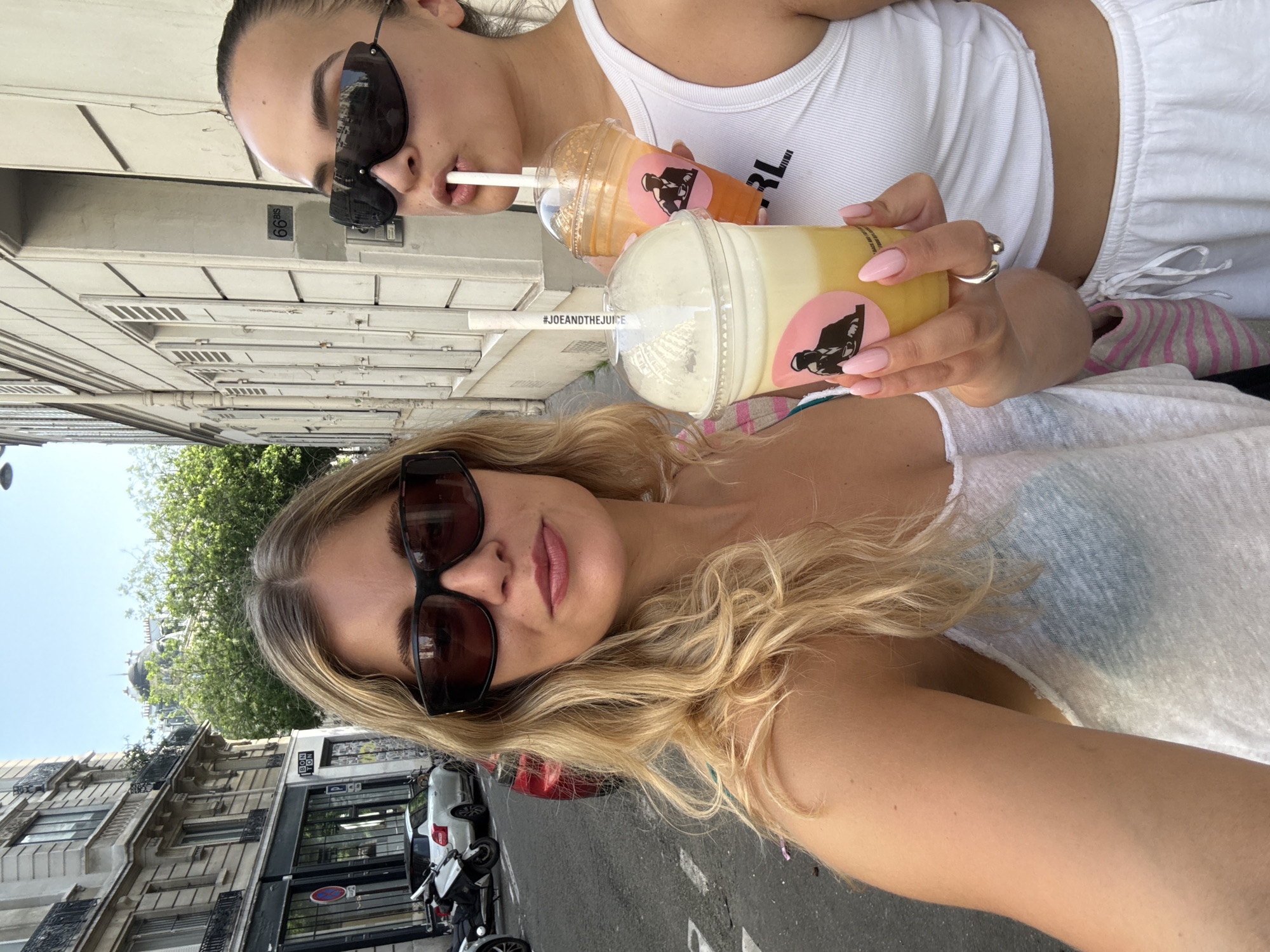 Two women are captured in a selfie, each holding a Joe & The Juice beverage. The woman closer to the camera has blonde, wavy hair and wears sunglasses and a white top. Her drink appears to be a light-colored fruit smoothie. The second woman, positioned behind the first, has dark hair pulled back and is also wearing sunglasses and a white tank top. She is drinking from a bright orange-colored drink. Both beverages bear the Joe & The Juice logo.
The location appears to be a street in Paris, France. Buildings with classic Parisian architecture are visible in the background. A motorcycle is parked on the side of the street, which has marked pedestrian crosswalk lines. The sky is clear and sunny, suggesting a daytime setting. The camera is angled slightly upwards, capturing the faces of the two women and the surrounding urban environment.
