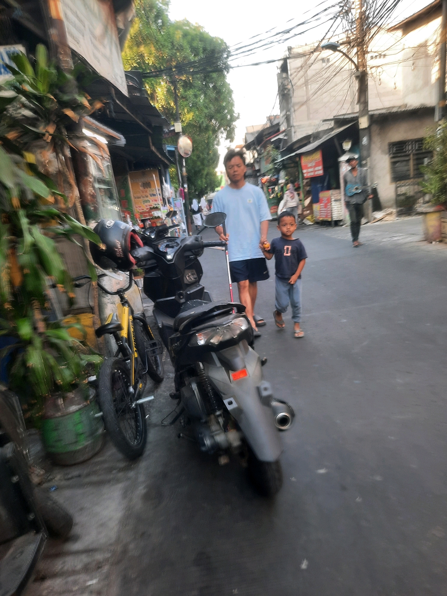 The image depicts a street scene in North Jakarta, Indonesia, characterized by its urban setting with a mix of shops and residential buildings. The foreground features several parked motorcycles, including a black scooter and a yellow bicycle. A person, likely an adult male wearing a light blue t-shirt and dark shorts, is walking on the right side of the frame, holding a white cane. Beside him, a young boy in a dark t-shirt and grey pants is walking, holding an orange object. In the background, more buildings, utility wires, and vegetation are visible, suggesting a busy street. The lighting indicates it is likely daytime, possibly late afternoon, given the shadows. The street appears to be paved, and the general atmosphere is that of a typical urban environment in Indonesia.