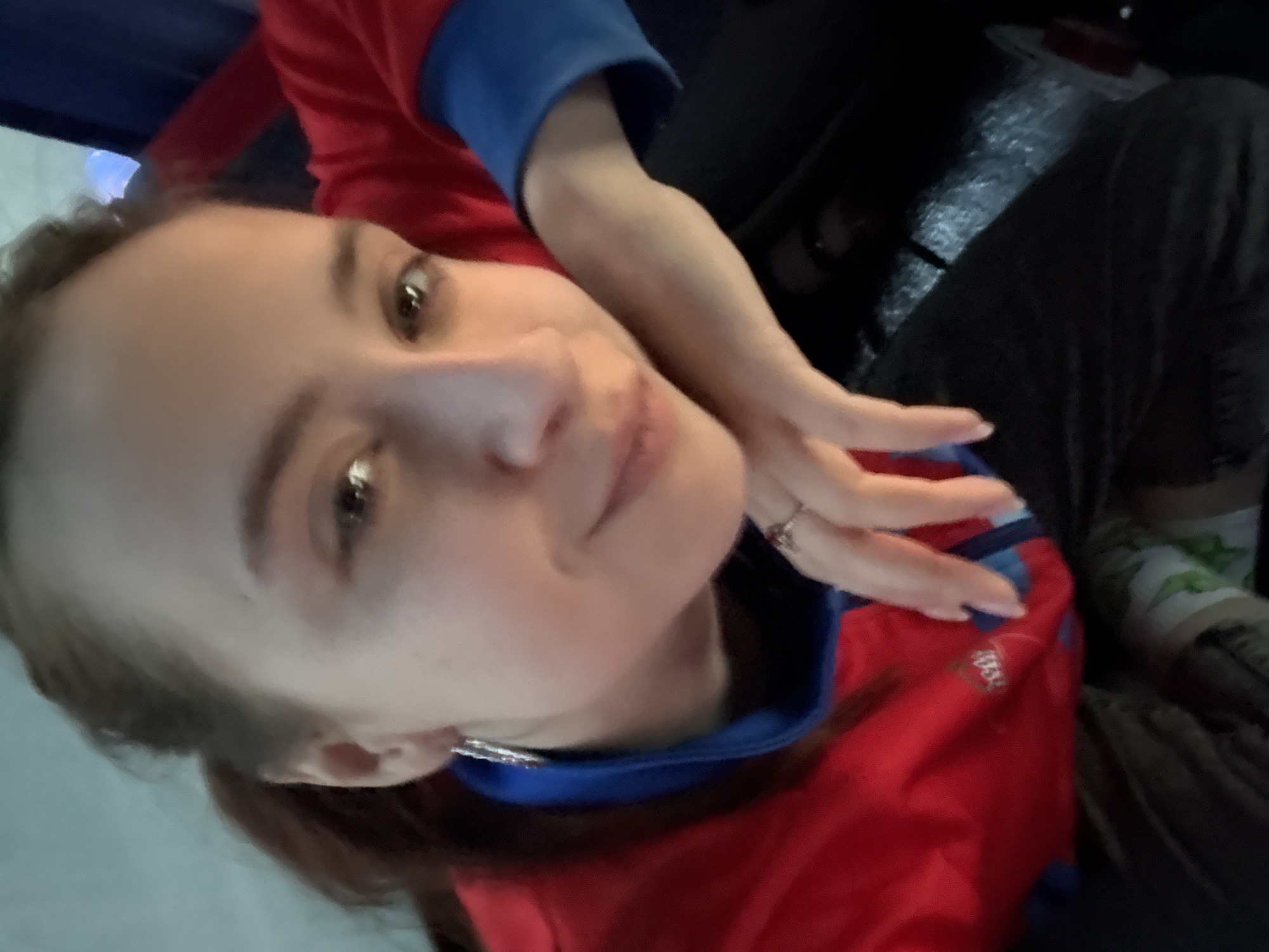 A woman is visible in the image, taking a selfie. She has fair skin, light brown hair pulled back, and is wearing a red shirt with blue accents. She is holding her hand near her face, and a ring is visible on one of her fingers. The background appears dark and out of focus, though a part of black jeans can be seen, along with a partially obscured leg possibly wrapped in a white bandage with what appears to be green shapes. It is difficult to determine the exact location without more context. Based on the query referencing "Permyakova, Russia," it could be inferred that she is in Permyakova, Russia, but this cannot be confirmed from the image alone.