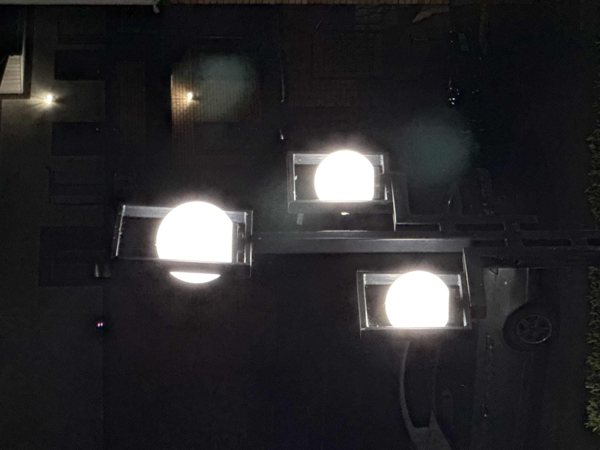 This is an overhead, low-light photograph taken at night, likely in an outdoor setting with artificial lighting. The scene appears to be a paved area, possibly a yard or a courtyard, adjacent to a building. Dominating the image are three large, spherical, white lights, each encased in a dark, rectangular fixture. These lights are bright and emit a significant glow, creating halos around them and illuminating the immediate surroundings with a cool white light.
To the left of the frame, a portion of a building is visible with a white siding. Further back on the left, another smaller, square-shaped light fixture is mounted on the building's facade, emitting a warm yellow light. In the upper right portion of the image, there's a hint of greenery, suggesting a garden or natural foliage bordering the paved area.
In the lower right of the frame, a dark, large object is partially visible, with a round, spoked element that resembles a wheel. This could indicate a vehicle or some other large equipment present in the scene. There are also some indistinct dark shapes and structures in the background, suggesting the presence of furniture or other outdoor elements.
The overall impression is one of a well-lit outdoor space at night, possibly designed for evening use or security. The presence of the building, paved area, and greenery points to a residential or commercial property. The image lacks any discernible human figures. No text is visible within the frame. The time of day is clearly night due to the artificial lighting and the dark background.