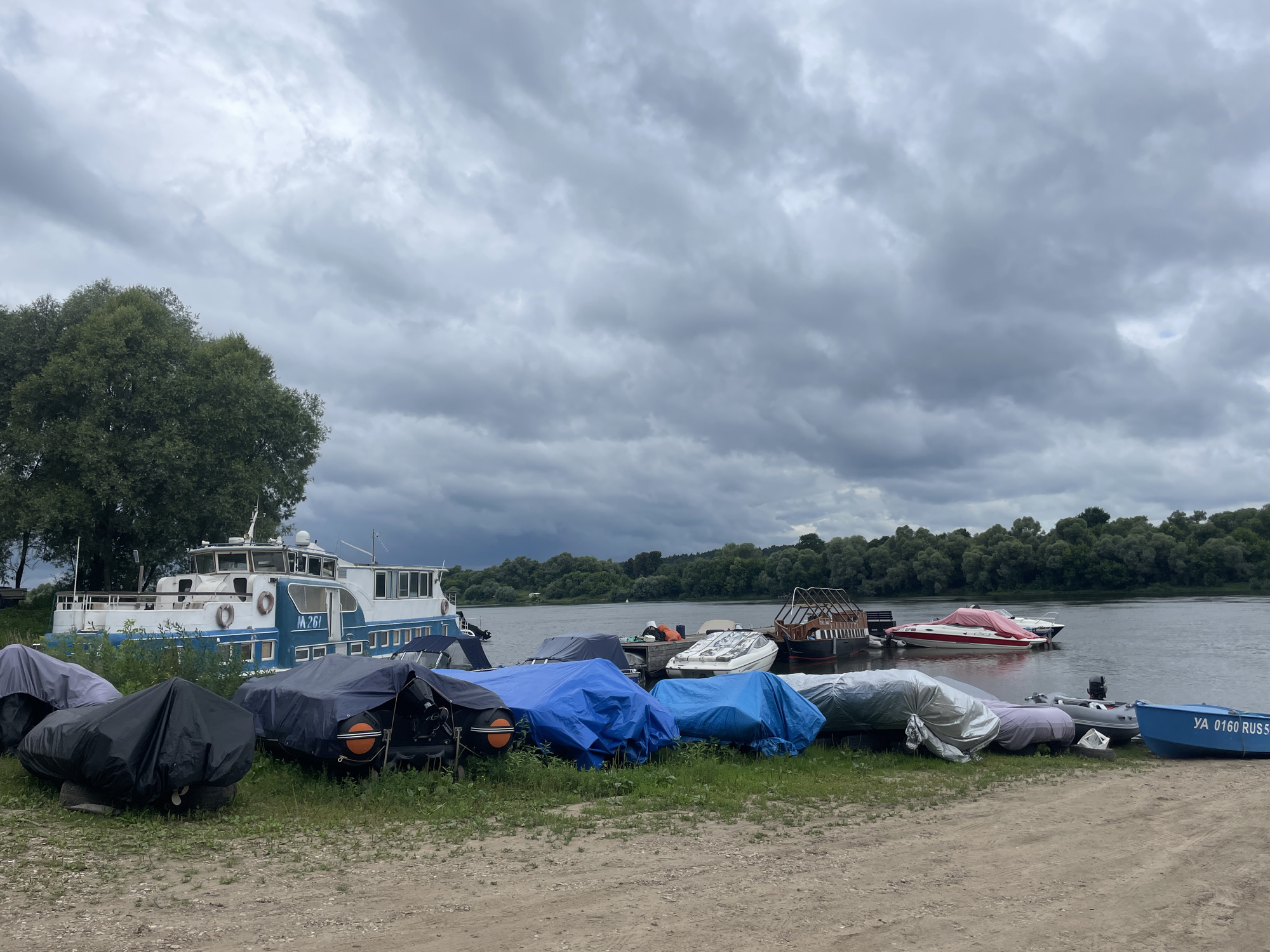 The image captures a riverside scene in Pushchino, Russia, under a cloudy sky. A large white and blue passenger boat, identified by the marking "M 261" on its side, is docked on the left. In front of it, on the grassy bank, numerous smaller boats are covered with tarps in various colors, including black, gray, and blue. Some of these covered boats appear to be speedboats or dinghies. Further out on the water, several other boats are visible, including a small white speedboat with a red cover, and a boat with a wooden superstructure. The opposite bank of the river is lined with a dense forest. The foreground consists of a sandy and gravelly area leading down to the grass. There are no people visible in the image.