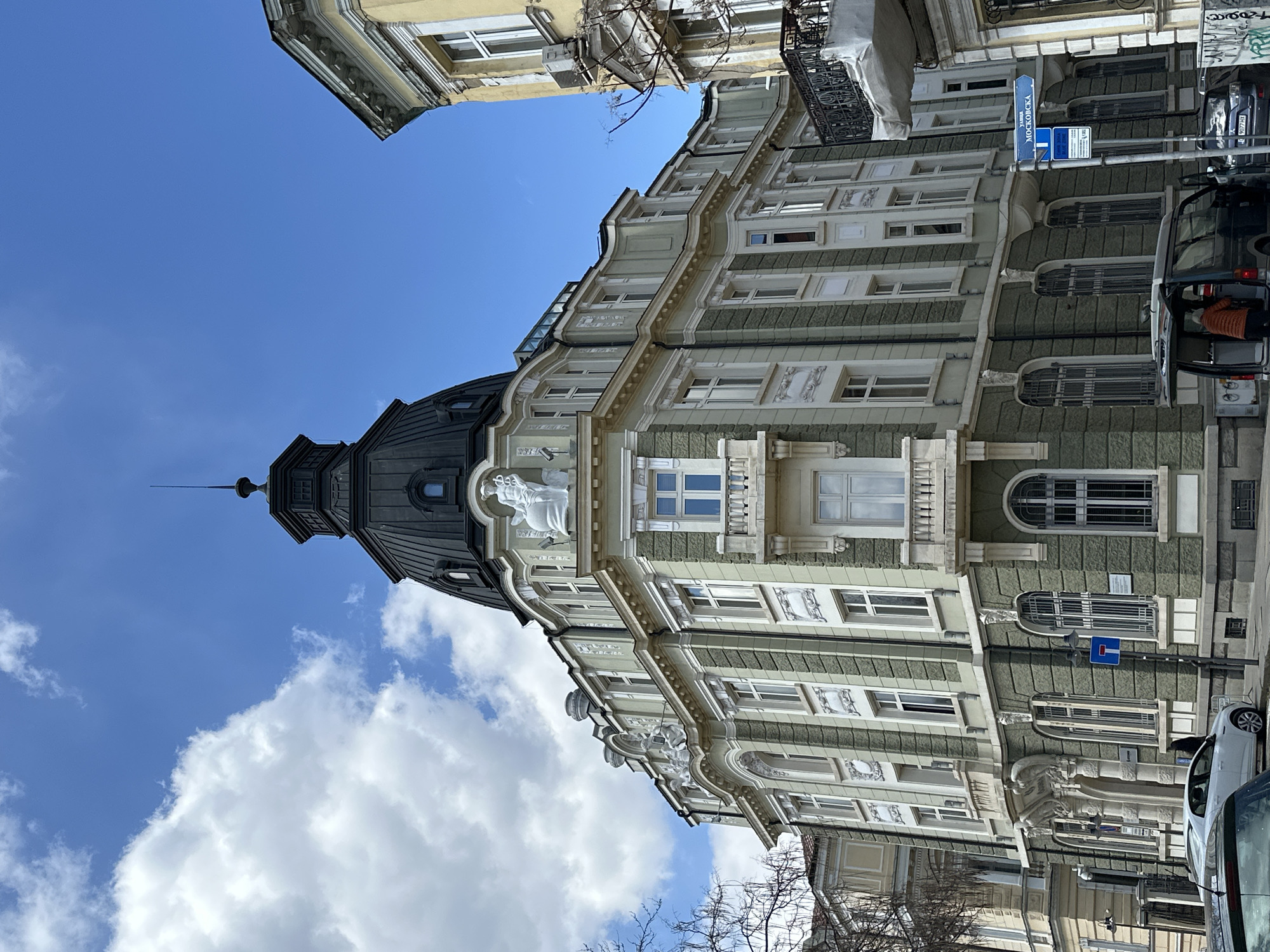 The image captures a view of a building in Sofia, Bulgaria, as indicated by the Cyrillic script on a street sign that says "Московска" ("Moskovska" in English transliteration). The architectural style of the building is classic and features elements like ornate window frames, statues, and a dark-colored dome atop a corner section of the building. The color scheme is mostly pale green and white, with the windows and decorations being in white.
The sky is visible in the background, showing a mix of blue sky and scattered white clouds. There is also a section of another building visible at the top left corner of the frame, indicating a dense urban environment.
Vehicles, including a white car and a dark van with the passenger side door open, are parked alongside the building.