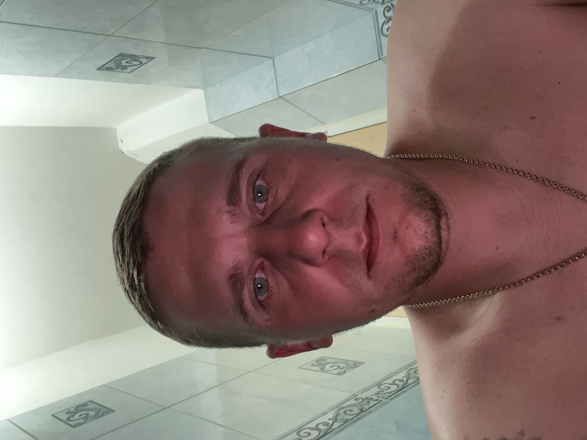 The image is a close-up, low-angle selfie of a man in what appears to be a bathroom. The man has short brown hair, blue eyes, and is wearing a gold chain. His skin appears flushed, possibly from heat or lighting. The background shows tiled walls, with a distinct pattern on the lower portion of the wall. The lighting is bright and direct, casting a reddish hue on the man's face. There are no identifiable landmarks or specific indicators of the city of Minsk, Belarus.