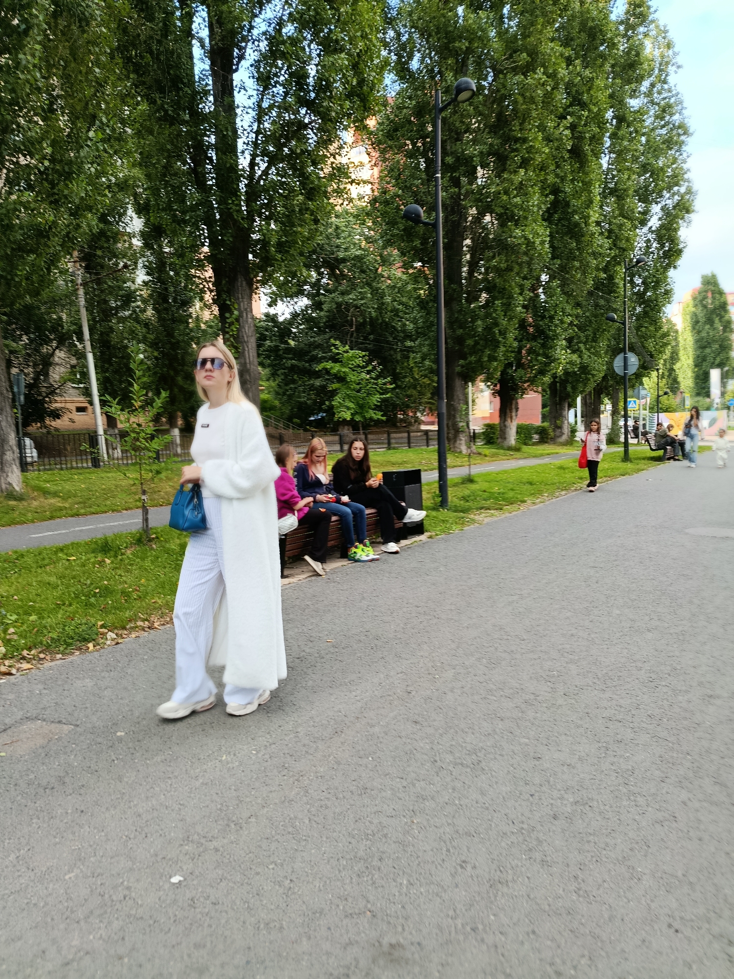 A woman in a long white cardigan and striped pants walks on a paved path in a park. She holds a blue handbag. In the background, a group of people sit on a bench, and another person walks in the distance. Tall trees line the path, and buildings are visible behind them. The scene appears to be in the city of Saratov, Russia.