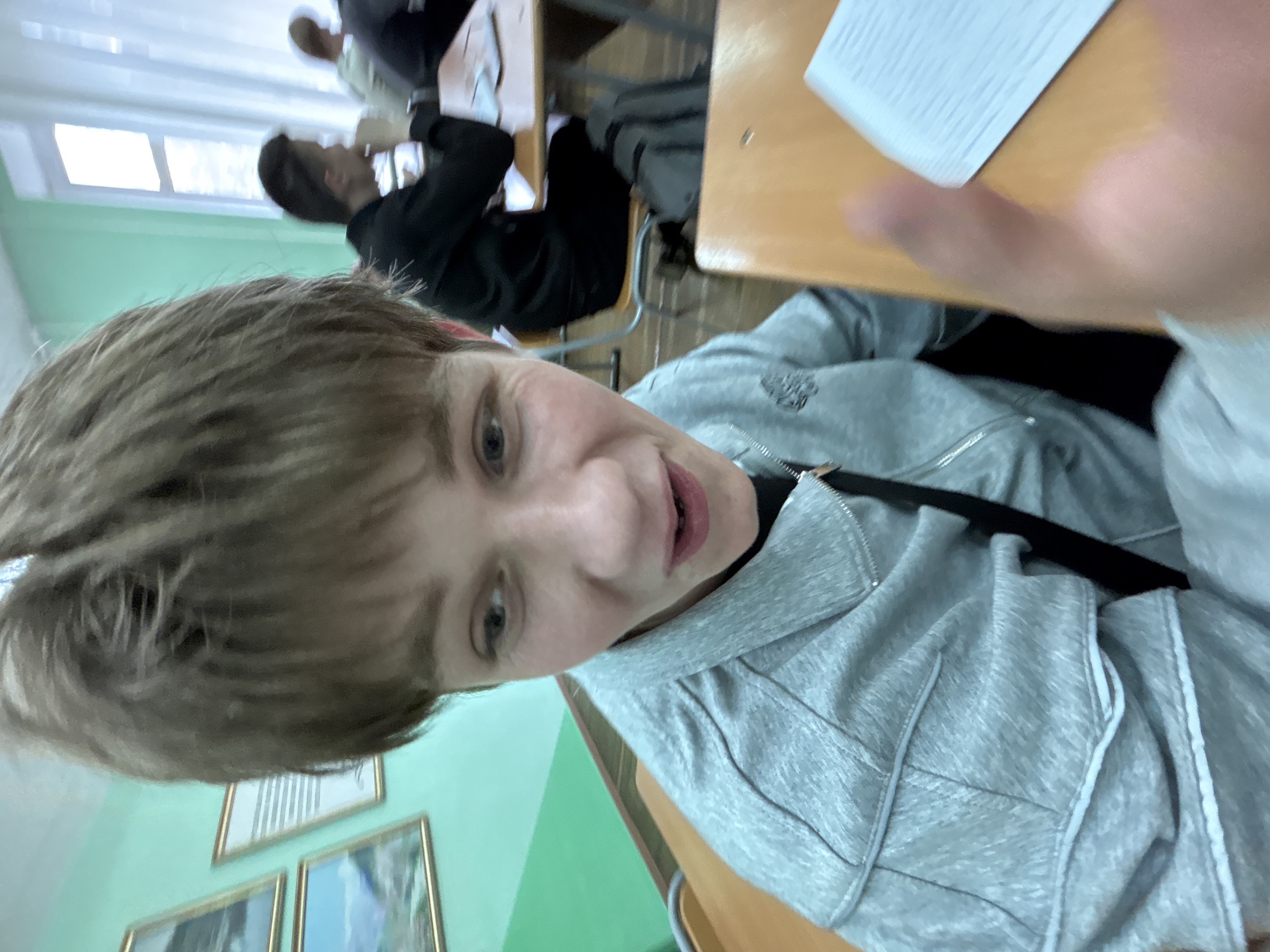 A candid photograph captures a young person in a classroom setting. The main subject, a light-haired individual, is in the foreground, looking directly at the camera with a casual, almost smirking expression. They are wearing a grey hooded zip-up jacket over a dark top. The upper part of another person's hand is visible in the top right, holding a white piece of paper or card.
In the mid-ground and background, other students are visible seated at light-colored wooden desks. One student with dark hair, wearing a black top, is clearly discernible further back. The classroom has light green walls and large windows on the left, which let in natural light. Several framed pictures or posters are hanging on the wall below the windows. The scene depicts an informal moment, possibly during a class break or a relaxed segment of a lesson. No legible text is visible in the image.