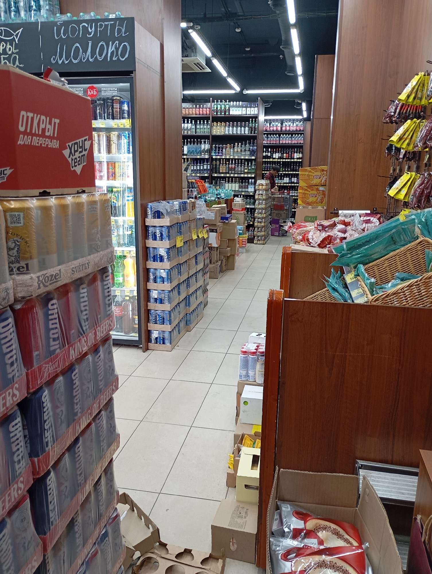 Aisle of a retail store, likely a supermarket or convenience store, with a selection of beverages and packaged goods. On the left, a display of stacked cans wrapped in plastic showcases brands like "Kozel" and "King of Beers." Behind this, a refrigerated display case contains various bottled drinks. Further back, shelves are stocked with a wide assortment of alcoholic beverages, including bottles of spirits and wine. The store interior features tiled flooring, wooden shelving units, and overhead fluorescent lighting. A sign on the left reads "ЙОГУРТЫ МОЛОКО" (Yogurts Milk) in Cyrillic script. The overall scene depicts a typical retail environment focused on beverage sales. The image does not provide specific location details for Moscow, Russia.