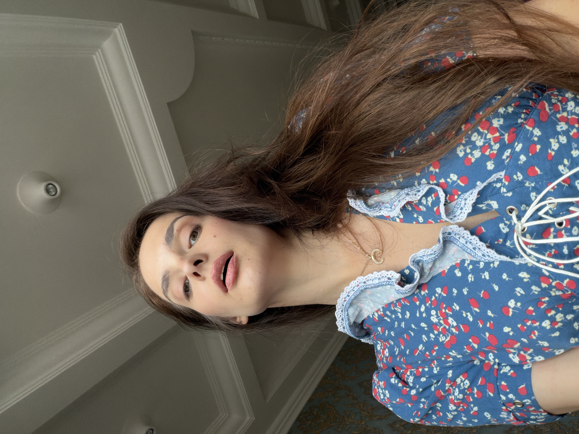 A woman with long, dark brown hair and brown eyes is shown from a low-angle perspective, looking upwards towards the camera. She is wearing a blue dress with a red and white floral pattern and white lace trim. The dress has a lace-up detail at the neckline. A delicate gold necklace with a "C" pendant is visible. Her lips are painted with a pink lipstick. The background appears to be an indoor setting, with white decorative molding on the ceiling and a wall with a subtle patterned wallpaper. There is a light fixture visible on the ceiling. The lighting suggests it could be daytime, possibly with natural light coming from a window, indicated by the shadows and highlights on the ceiling. The woman's expression is neutral. The image appears to be a selfie taken indoors.