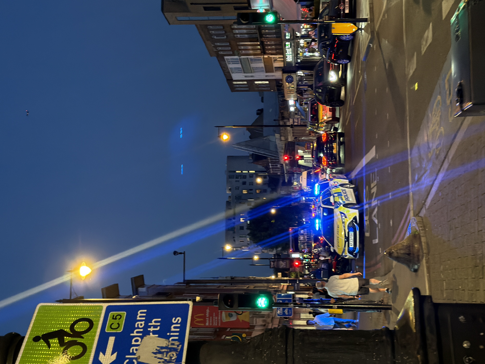 The image depicts a street scene in Clapham North, London, United Kingdom, at dusk or night. A police car with flashing blue lights is visible in the street, suggesting an incident or presence of law enforcement. Several other vehicles, including cars and a taxi, are also present, indicating a busy urban environment. Pedestrians are walking along the sidewalk. Street lighting is prominent, illuminating the scene, and traffic signals are in operation. Street signs provide directions for cyclists and vehicular traffic. Buildings line the street, and a McDonald's advertisement is visible. The overall impression is that of an active city street with a potential police intervention.