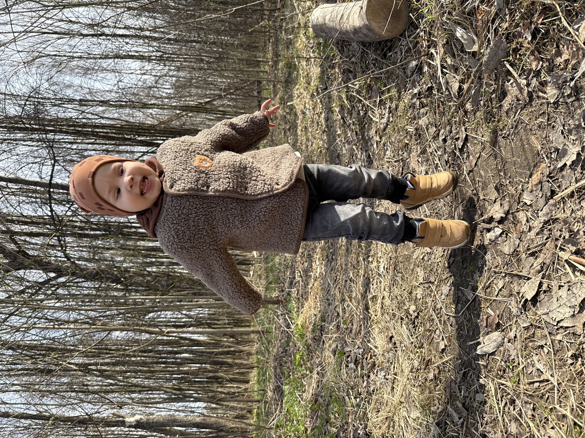 A full-shot portrait shows a young child standing upright in a wooded area. The child, likely a toddler or pre-schooler, faces the camera with a wide, joyful smile, revealing their teeth. They are dressed in a brown knitted beanie, a warm, fuzzy brown jacket (appearing to be sherpa material) featuring a small, circular patch on the left chest, dark grey pants, and tan-colored boots. Their right arm is slightly raised with the palm open, suggesting a wave or friendly gesture.
The scene is set in a forest or woodland. The ground is covered with an abundance of dry leaves, twigs, and sparse patches of light-colored, dry grass. In the background, numerous deciduous trees stand bare, their branches reaching towards a clear, bright sky, indicating late winter or early spring before foliage has appeared. A cut log lies on the ground to the right of the child. The time of day is sunny daytime, and the child's warm clothing suggests a cool temperature. No text is visible in the image.