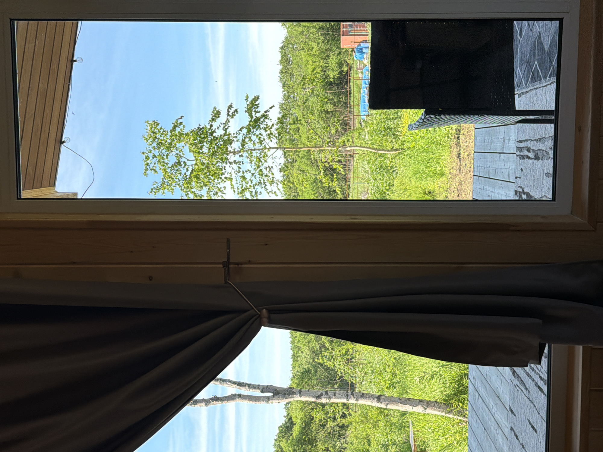 The image is a view through a window of a wooden structure looking out onto a rural landscape on a sunny day. A dark gray curtain is pulled to the left, revealing a wooden deck in the foreground. Beyond the deck, there is lush green vegetation, including trees and tall grass. In the middle ground, a thin, bare tree trunk stands to the left of a larger, leafy tree. Further back, there's a fence line, a blue tarp, some blue barrels, and a red structure. The sky is clear blue with a few faint clouds and a small airplane visible. The wooden structure appears to be a cabin or room with wooden walls and a wooden ceiling with string lights. The scene is consistent with a location in a natural, possibly recreational area, such as the outskirts of a town like Новый, Russia, where such wooden dwellings and open natural spaces are common. There are no people visible in the image.