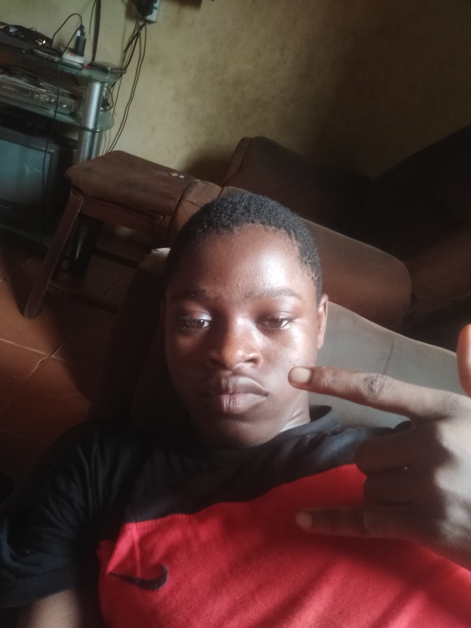 A young person, likely male, with short, dark hair and dark skin, is depicted in a close-up, slightly angled indoor photograph, possibly a selfie. The individual is reclining or sitting back on what appears to be a brown upholstered couch or chair, looking directly at the camera with a neutral to slightly serious expression. There is a noticeable sheen on the forehead.
The person is wearing a black t-shirt with a large red section across the chest, featuring a black Nike swoosh logo. The right hand (viewer's left) is raised, forming a gesture with the index finger extended forward and the pinky finger also extended, while the other fingers are curled.
The background on the left reveals an indoor setting with various household items. A dark television screen is visible at the bottom left, topped by a glass-shelved unit holding electronic equipment like a stereo or DVD player, along with tangled cables and chargers. A brown wooden chair or furniture leg is also present. The wall behind is a light brown or beige color with some texture. The right side of the background is largely obscured by the dark couch. The scene suggests a casual indoor environment, likely a home in Shangisha, Nigeria.