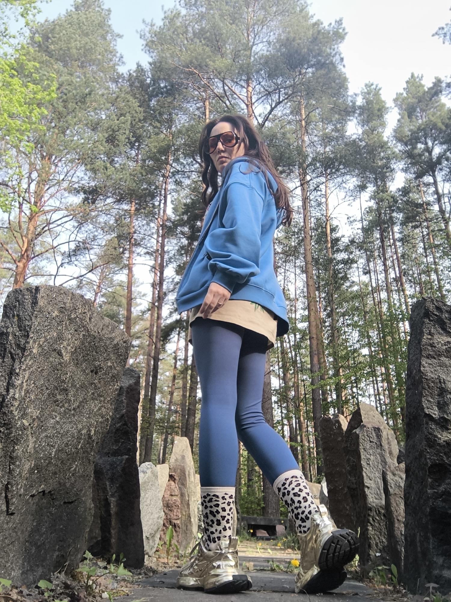 A low-angle, medium shot depicts a woman standing in the Memorial to the Victims of Nazism, located in Riga, Latvia. She is angled slightly to the right, looking over her left shoulder towards the camera. She wears tinted sunglasses, a light blue hooded sweatshirt, a khaki shirt underneath, blue leggings, patterned socks, and metallic gold sneakers. Tall pine trees and granite stones form the backdrop. A patch of green leaves is visible in the upper left corner. The sky is visible through the trees.