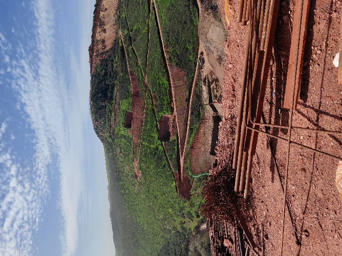 The image depicts an expansive, open-pit mining or construction site within a hilly, semi-arid landscape in Fdalate, Morocco. The foreground is dominated by a reddish-brown earth slope, where numerous rust-colored metal elements are scattered. These include bundles of rebar, individual rebar pieces, elongated cylindrical metal pipes, and flat metal beams. A large, dense pile of tangled rebar scraps is visible on the left.
The midground features a series of winding, unpaved reddish-brown roads or terraces cut into steep, verdant hillsides covered in green scrub vegetation. Several rectangular, dark openings or structures are embedded into these excavated hillsides, appearing as voids or reinforced entrances. Further down and right, a large, cleared area of lighter-colored earth and debris is visible at the base of the hills, indicative of a material processing or dumping zone. No individuals are discernible in the scene.
The background shows a distant, higher peak, partially covered in green vegetation and partly composed of exposed reddish-brown rock and earth. A cluster of buildings or structures is visible on the summit of this peak. The sky above is clear blue with scattered white, cumulus clouds. The scene is illuminated by strong sunlight, casting distinct shadows.