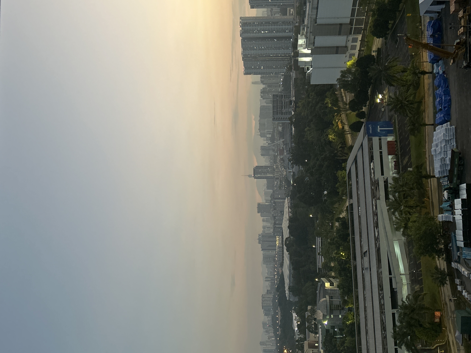 This is a wide-angle, high-angle shot of a city skyline at dawn, likely Kuala Lumpur, Malaysia, given the presence of distinctive buildings like the Petronas Twin Towers (visible in the distance) and the distinctive architectural style of the surrounding cityscape.
The sky is a pale gradient, transitioning from a soft blue at the top to a warm, peachy hue near the horizon, indicating sunrise. Wispy clouds are scattered across the sky.
In the mid-ground, a dense urban area is visible, characterized by numerous high-rise buildings of varying heights and designs. A prominent stadium with a distinctive white, curved roof is situated within this urban landscape. Lush green trees form a dense canopy throughout this area, suggesting parks or landscaped areas interspersed with buildings.
In the foreground, a more industrial or commercial zone is depicted. A long, white, multi-story building with a flat, covered roof stretches across the frame. This building appears to be a parking garage or a similar structure. Below and in front of this structure, a parking lot is visible with scattered palm trees and some smaller trees.
To the right of the foreground building, a large, modern white building complex with several rectangular sections is present. Further in the foreground, on the ground, several stacks of white materials, possibly construction materials or supplies, are arranged. Blue tarpaulins cover other materials. A construction crane, with its boom extended, is positioned among the covered materials. A dark green truck is also visible to the left of the white stacks.
The lighting suggests early morning. The overall impression is one of a bustling city awakening, with residential and commercial areas coexisting with green spaces. No people are clearly discernible in the image.