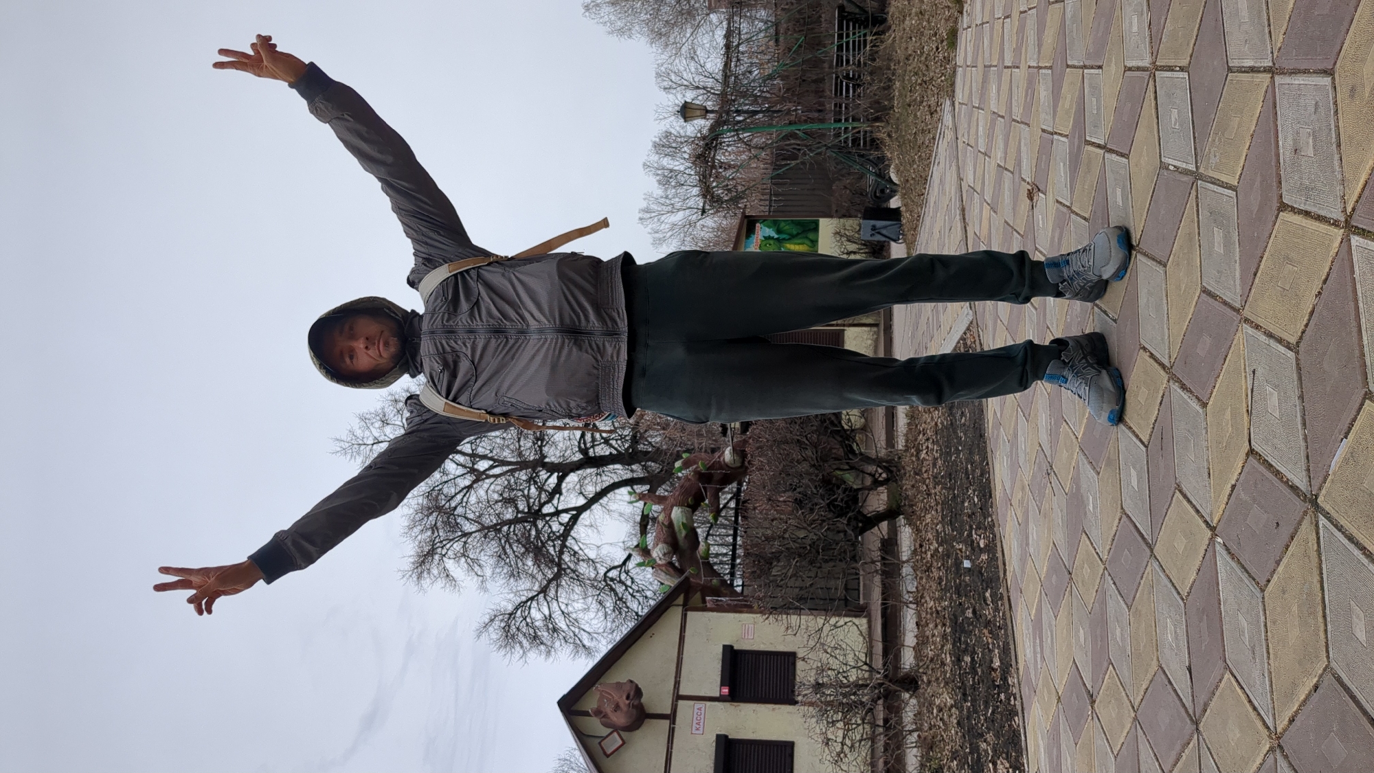 A man stands on a patterned brick walkway in what appears to be a park or outdoor area in Ulyanovsk, Russia. He is wearing a grey jacket with a hood, dark trousers, and grey running shoes with blue accents. He has his arms outstretched, making a peace sign with his right hand. Behind him is a building with the word "KACCA" displayed and what appears to be a large, sculpted head. There are bare trees in the background and a cloudy sky overhead. In the far background there is a green metal playground structure.