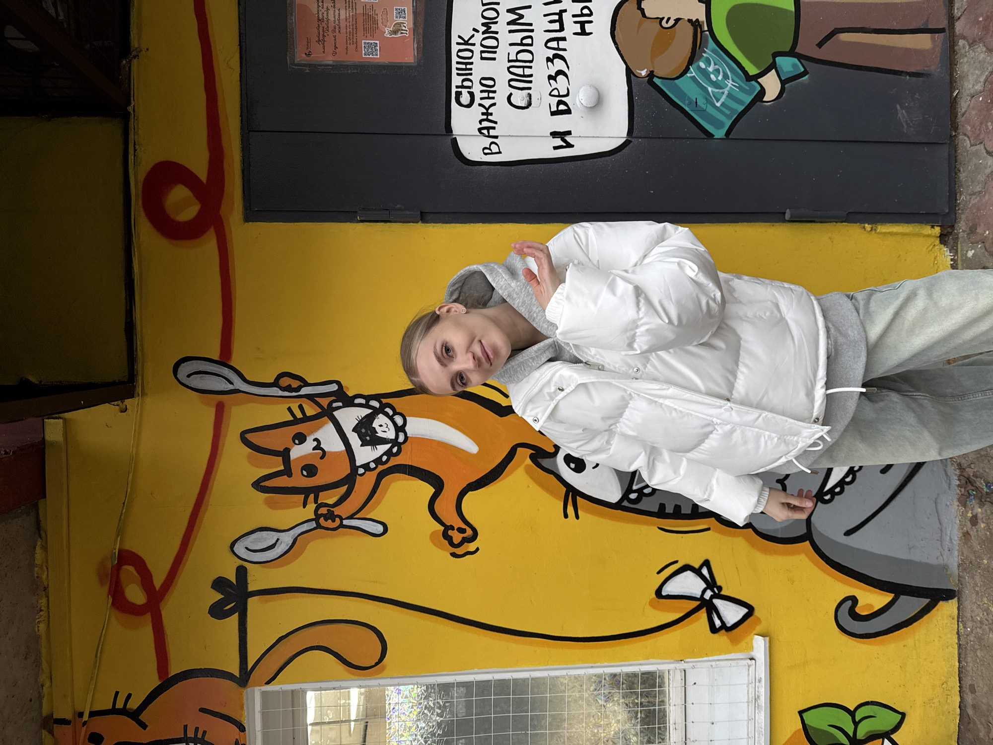 The image shows a woman standing next to a colorful mural on a building in Izhevsk, Russia. The mural features cartoonish cats and mice, some holding spoons. Above the mural is a dark painted door with a speech bubble containing Russian text. The woman is fair-skinned, has blonde hair, and is wearing a white puffer jacket over a grey hooded sweatshirt and light blue jeans. She is posing with one hand raised. The surrounding area appears to be urban, with a mix of painted walls and what looks like street paving. The location is Izhevsk, Russia based on the OCR content of the image, which translates to indicate support for animal shelters in the city.