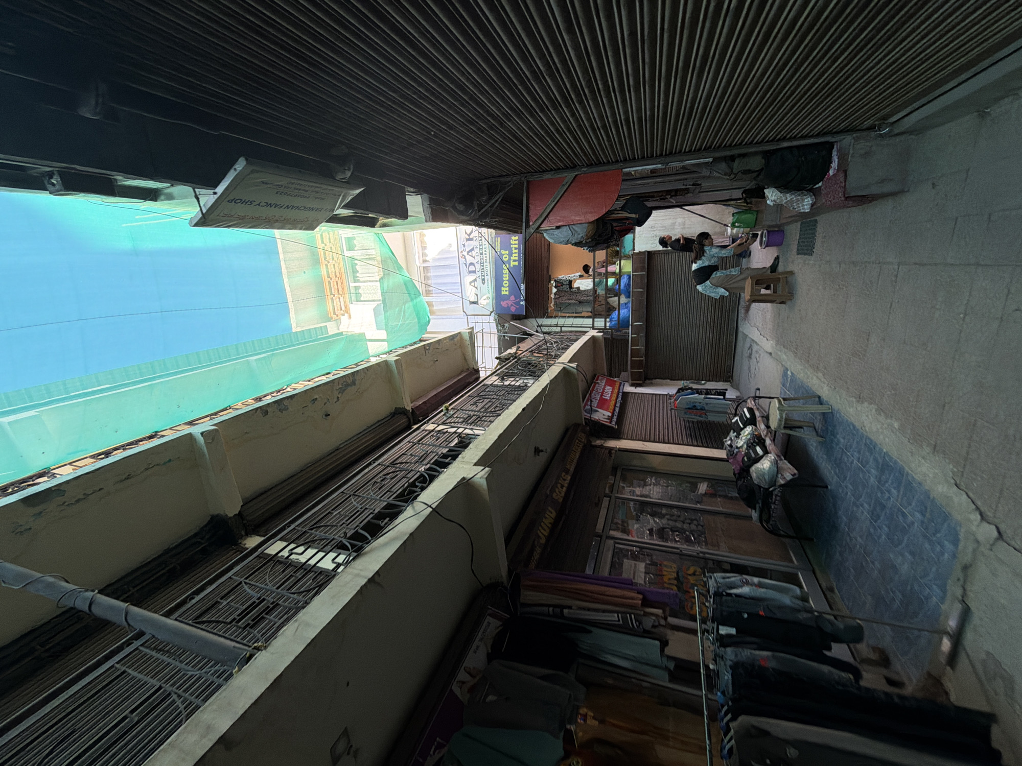The image depicts a narrow alleyway in Leh, India, likely in a market area. On the left, a multi-story building features balconies with metal railings and closed shutters on the lower floors, suggesting shops or residences. Clothes are displayed on racks and draped over a small table. The ground floor of this building has shop fronts with glass windows.
On the right side of the alley, a building is covered with corrugated metal shutters. A green tarpaulin is stretched overhead, covering parts of the alley and buildings. Several signs are visible: one reads "LABAK ACADEMY" and "TUTION CENTRE," another says "House of Thrift," and a third, on the far right, shows "SALOON" with some text below.
Two women are present in the alley. One woman, wearing a light-colored shirt and dark pants, is seen from the back, appearing to be cleaning with a green jug and a purple bucket. Another woman, wearing a dark top and patterned skirt, stands near her. A wooden stool is placed near the cleaning woman. Further into the alley, another person is partially visible.
The ground is paved with concrete. The overall impression is of a bustling, somewhat cramped commercial or residential street with shops and daily activities taking place. The bright blue sky is visible through the green tarpaulin at the top of the frame.