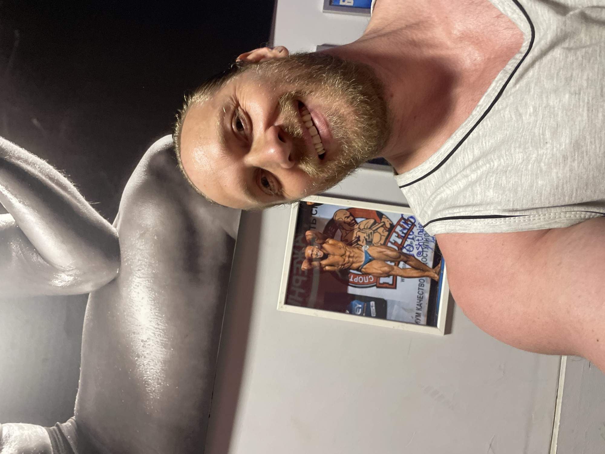 A man with a beard and light brown hair tied back in a ponytail takes a selfie in what appears to be a gym. He is wearing a grey tank top. In the background, there is a large black and white poster of a muscular person's back, and a framed picture of a bodybuilder in a blue posing trunk. The man is smiling at the camera. The environment suggests a bodybuilding or fitness-oriented establishment, possibly within Saint Petersburg, Russia, given the Cyrillic text visible on the framed picture.