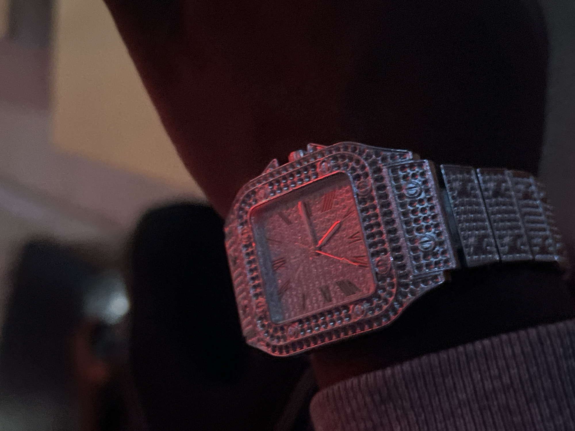A close-up shot focuses on a silver-colored wristwatch with a rectangular face, heavily embellished with what appears to be small, round stones, encircling the dial and the entire band. The watch is worn on a person's wrist, which is partially visible with a light gray or white, ribbed fabric cuff near the watch. The watch face features Roman numerals and hands, reflecting a red light. The overall scene has limited visible context, making it challenging to determine the location in Jalingo, Nigeria, without additional information.