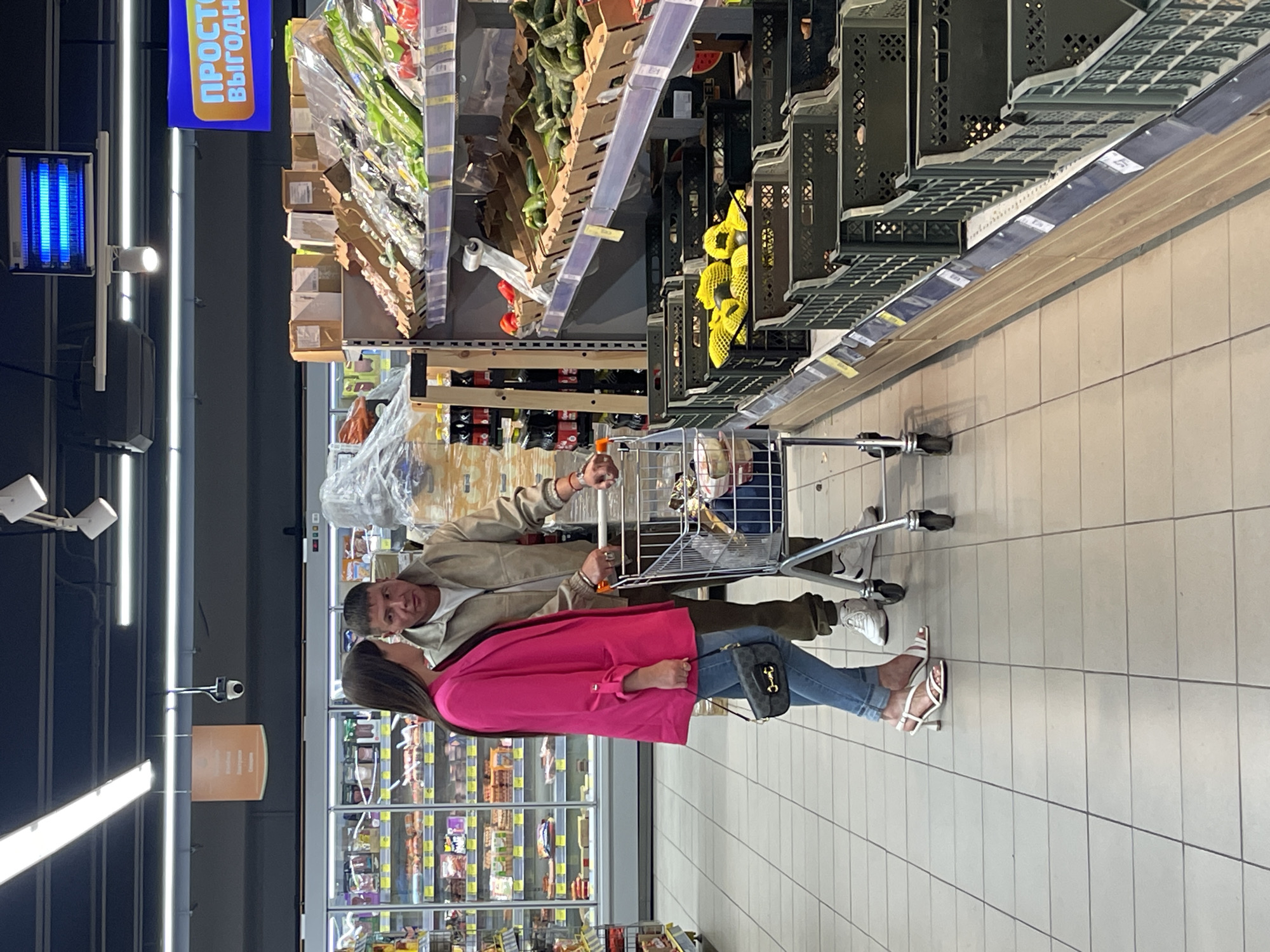 In Moscow, Russia, a couple shops in a supermarket. The woman, wearing a bright pink blazer, blue jeans, and white heeled sandals, stands on the left, holding a small black purse. The man, in a beige jacket and white sneakers, stands to her right, pushing a shopping cart. They are positioned between aisles of groceries, with refrigerators stocked with food items visible behind them. A shelving unit laden with produce, including cucumbers and bell peppers, is to their right. The floor is tiled, and overhead lighting illuminates the scene. A digital sign displaying Russian text is visible in the background.