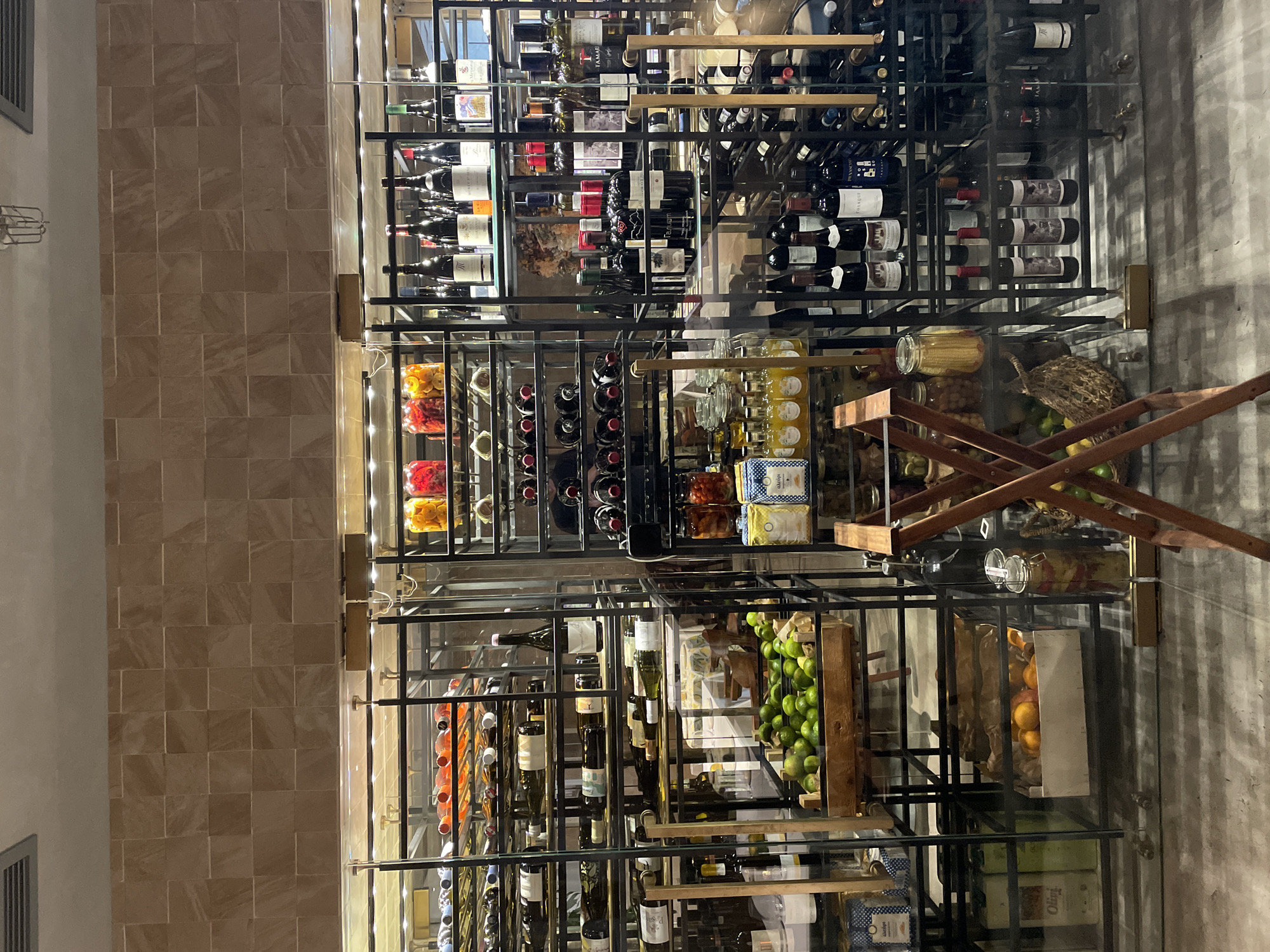 The image shows a multi-tiered shelving unit, likely inside a store or restaurant. The shelves are made of dark metal and glass, and they are filled with various items, including wine bottles, preserved foods in jars, lemons, limes, and oranges. There is also a wooden folding table or ladder partially visible in the center of the frame. The scene is well-lit. The floor appears to be concrete or tile. The walls in the background are covered with light brown rectangular tiles. The image does not provide any clear indication of its location in Saint Petersburg, Russia, or anywhere else.