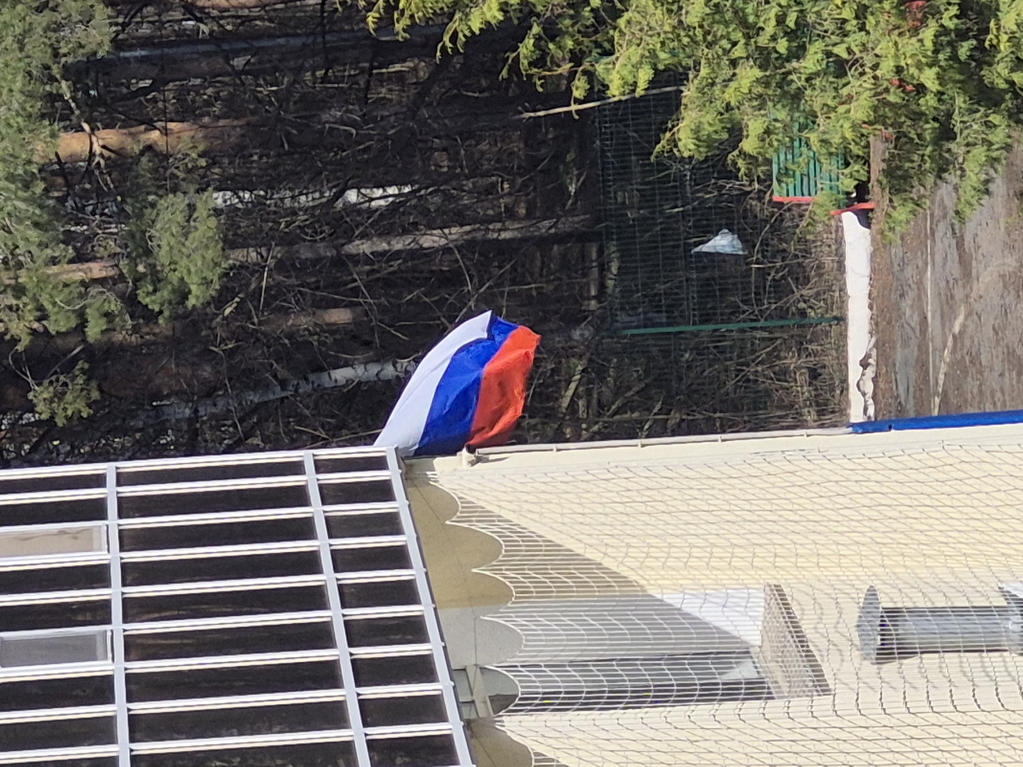 The image shows a Russian flag draped over the edge of a building in Chaikovsky, Russia. The building has a grid-like window structure on one side and a light-colored, possibly tiled, exterior with visible vents. The background consists of trees and a fence.