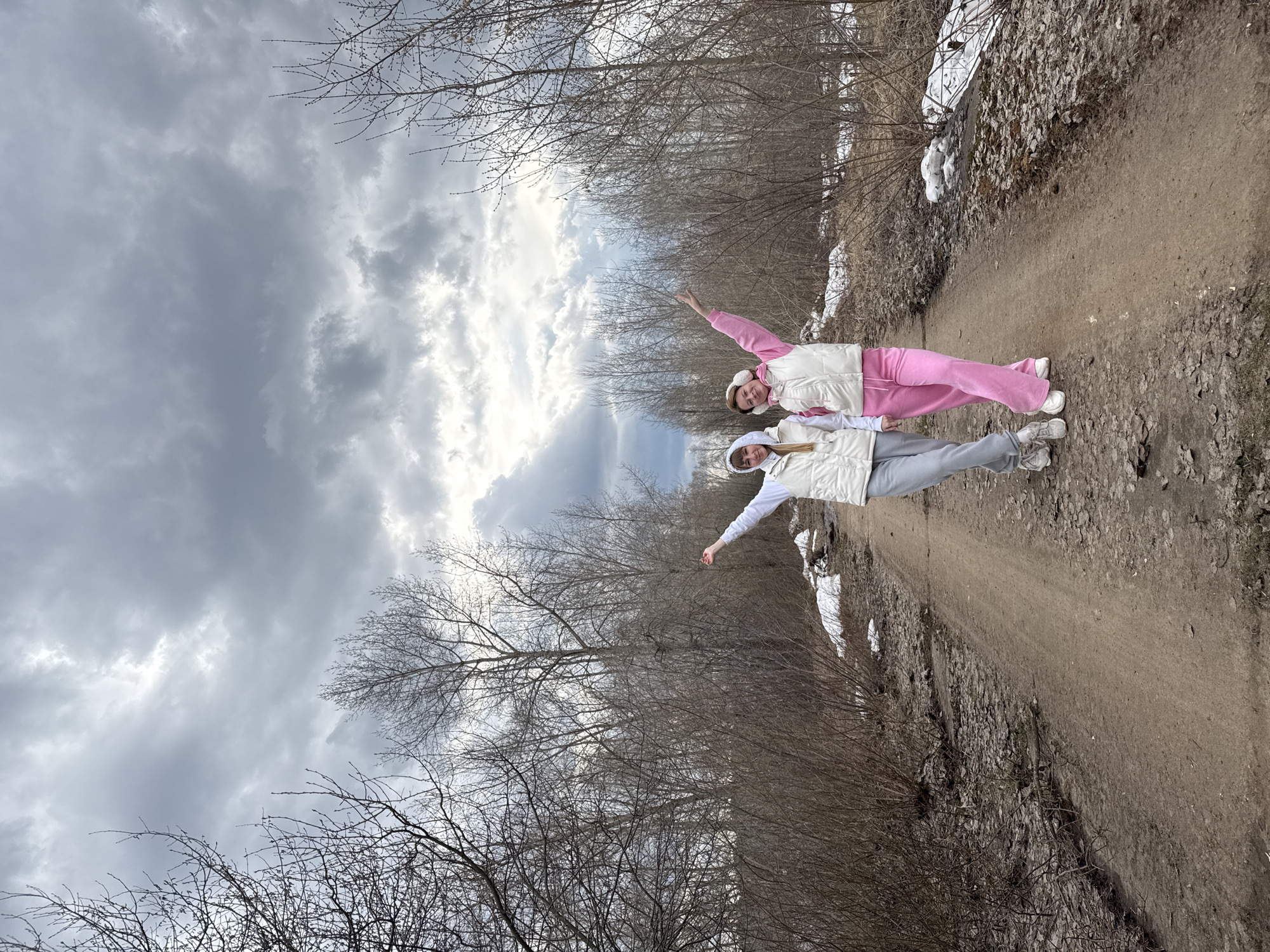 The image displays two young women posing playfully on a dirt path in Chaikovsky, Russia, during what appears to be late winter or early spring. The scene is characterized by bare trees and patches of snow, indicative of cold weather.
The woman positioned on the left wears a pink tracksuit with a white hooded vest and white sneakers. Her right arm is bent at the elbow, with her hand pointing upwards. Beside her, the second woman is dressed in a grey tracksuit, a matching white hooded vest, and grey sneakers. Her left arm is bent at the elbow, with her hand pointing outwards. Both women have hoodies under their vests, and the woman in pink is also wearing white earmuffs. Their relaxed and cheerful poses suggest a joyful outdoor outing.
They stand on a wide, unpaved dirt path, which is covered with dried leaves and small stones. Dense thickets of trees and shrubs, devoid of foliage, line both sides of the path. Several patches of snow are visible on the ground along the path's edges and among the vegetation. The sky above is heavily overcast with grey clouds, although some brighter sections suggest sunlight attempting to break through. The overall atmosphere is cool and natural. There is no visible text in the image.