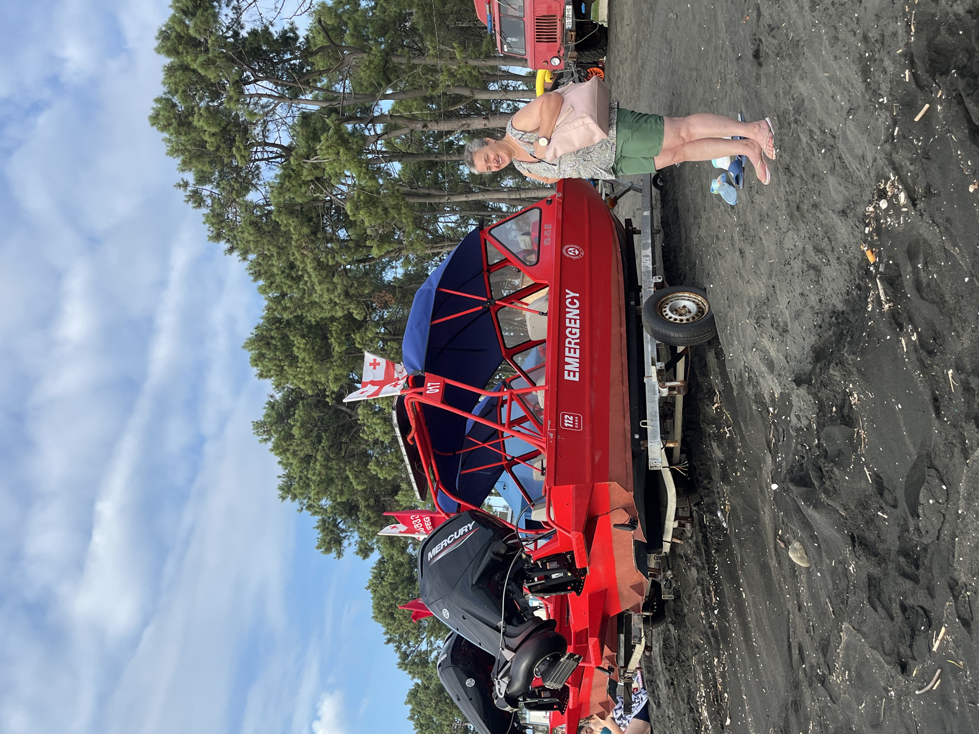 A woman stands on a black sand beach in Ureki, Georgia, next to a red emergency boat. The boat, mounted on a trailer, has two black Mercury outboard motors. The boat is labeled "EMERGENCY" and has a Georgian flag attached to a pole on its canopy. Behind the boat, a line of tall green trees forms a backdrop. To the right of the boat, a red truck is partially visible. The woman is wearing a patterned sleeveless shirt and green shorts, and she is carrying a light pink bag. She is wearing flip-flops. The sky above is partly cloudy.