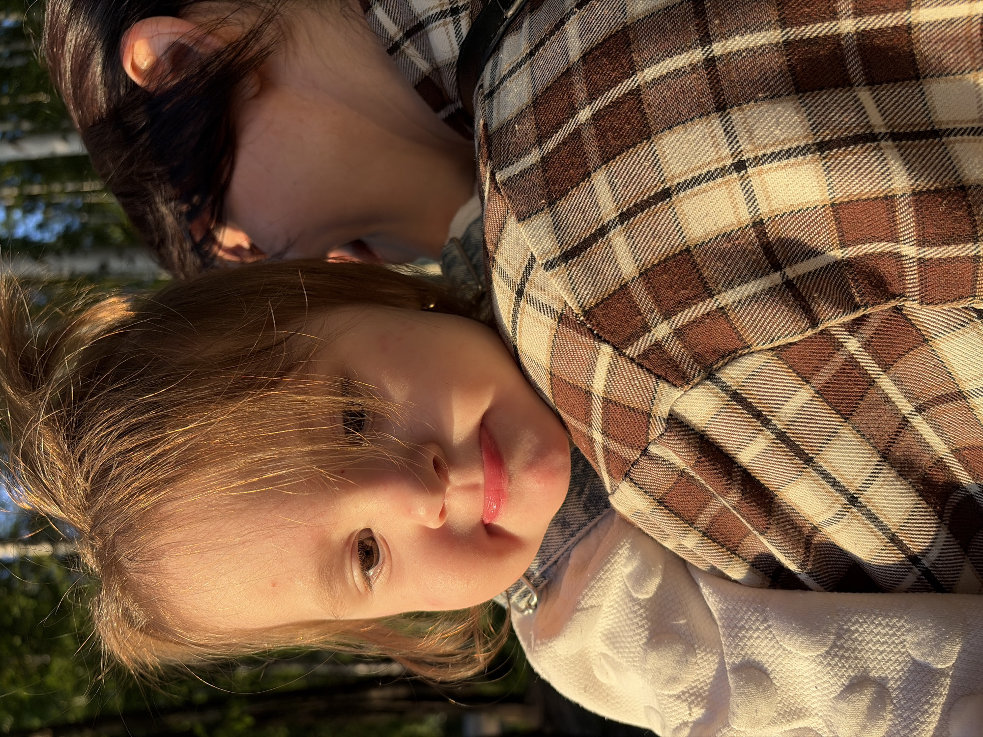 A close-up shot shows a young child being held by an adult in an outdoor setting. The child, with fair hair and rosy cheeks, looks directly at the camera with a slightly pursed mouth and wide eyes. The child is wearing denim overalls over a white, textured top with a dotted pattern. The adult, visible from behind, has dark hair and is wearing a brown and tan plaid shirt. The background suggests a natural environment with trees and sunlight filtering through. The image appears to be taken in a park or wooded area. There is no indication of the city of Chaikovsky, Russia, or any specific location within it.