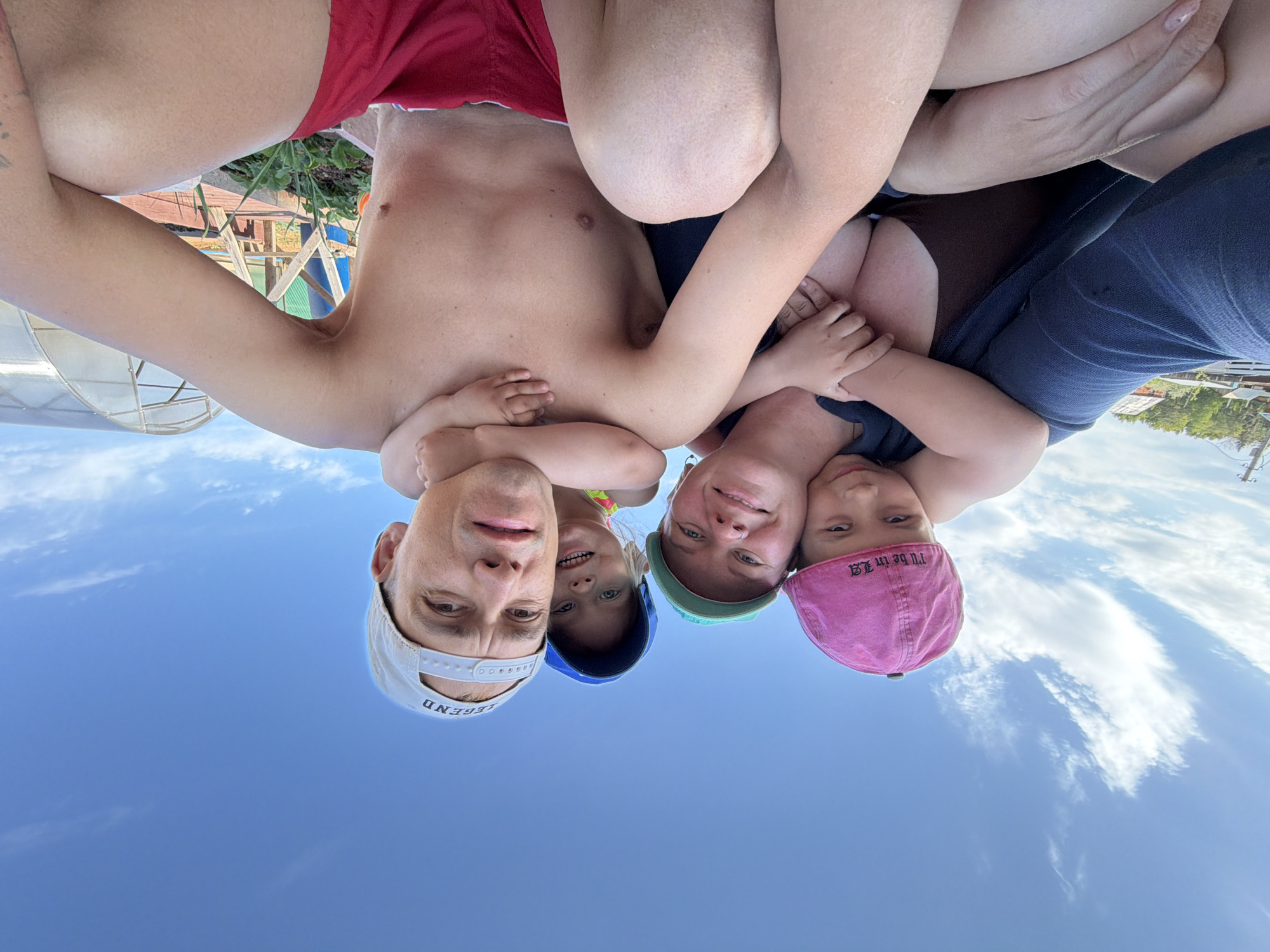 A selfie depicts a group of five people against a blue sky, appearing to be upside down. A man with short hair and wearing a white baseball cap embraces a woman and two children. The woman has short hair and wears a green cap. One child wears a blue cap, while the other wears a pink cap. The man is shirtless and wears red shorts. The photograph appears to be taken outdoors during the day. The background includes a few trees and structures, including a white awning. The location may be in or near Chaikovsky, Russia, based on the surrounding environment. The subjects are engaged in a close embrace.