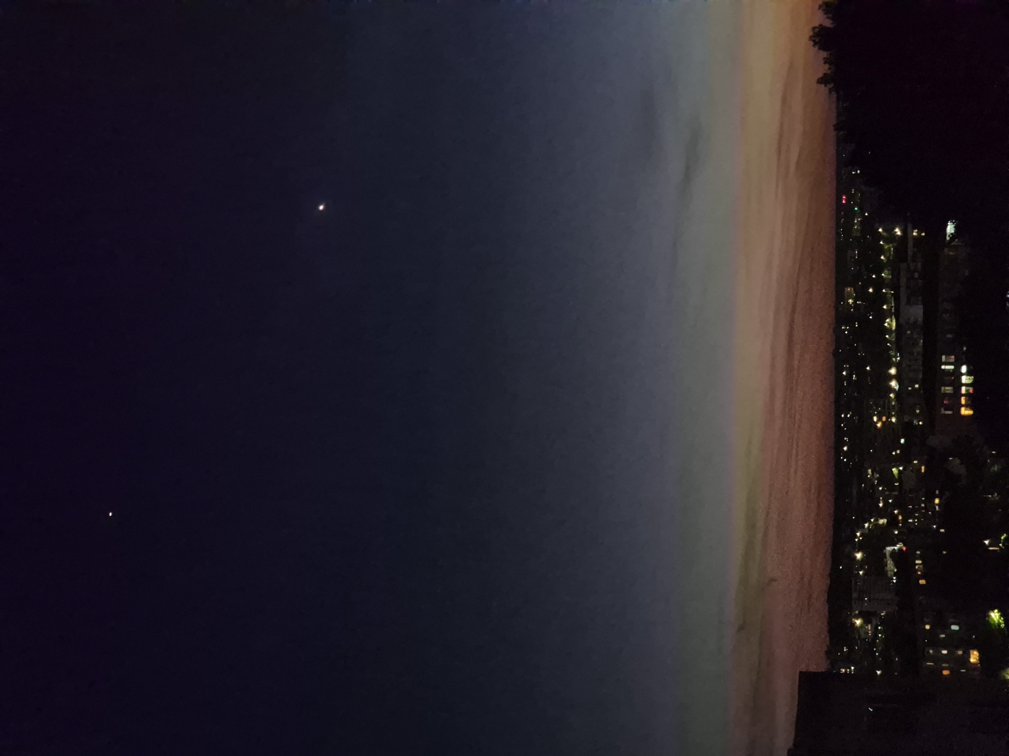 Here is a forensic description of the image:
The image appears to be a nighttime view of a cityscape meeting a body of water, possibly the ocean. The left side of the frame is dominated by a dark, twilight sky, with two discernible bright celestial objects. The right side of the image is occupied by the cityscape, indicated by numerous bright points representing artificial lights emanating from buildings and streets. The horizon line displays a gradient of color, transitioning from the dark sky into hues of orange and pink, possibly from a sunset. The city appears to be situated along the water's edge, with the horizon line acting as the boundary between the water and the sky. The location is Unknown, Unknown.